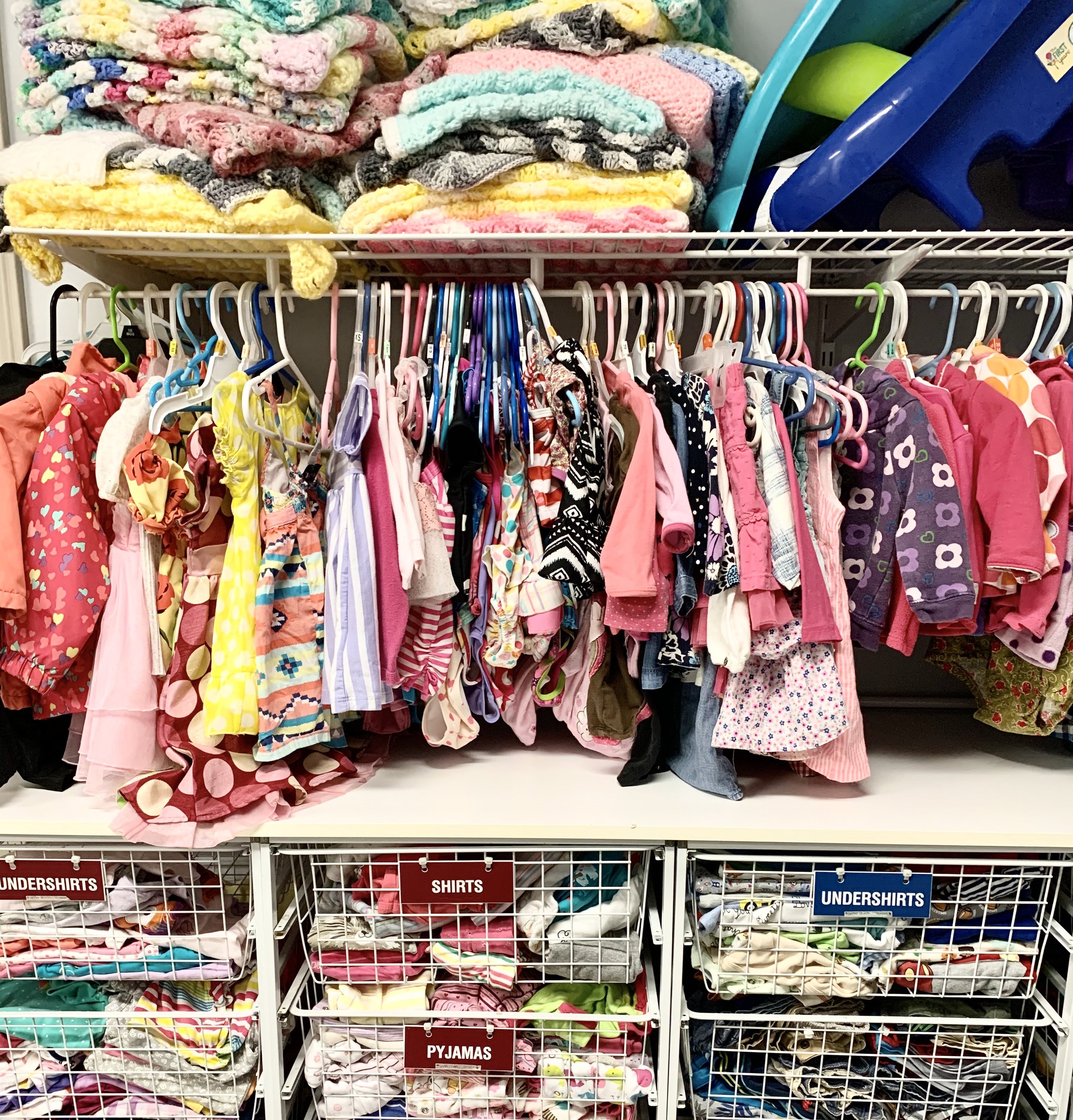 Children's clothing on hangers with shelves of folded clothes above and baskets of clothes below in a store. The clothing includes various dresses, shirts, and pajamas in bright colors and patterns.