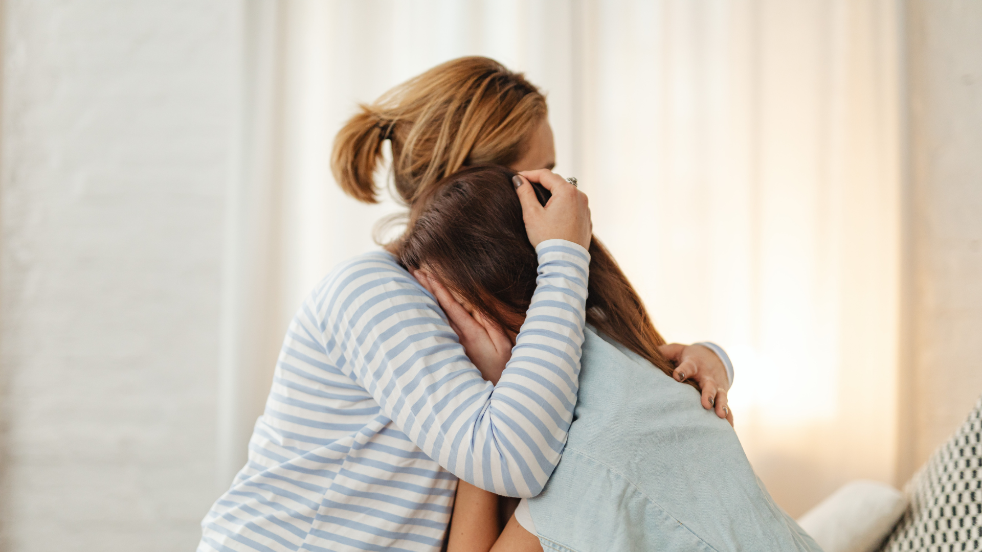 Two women hugging each other, one with light hair and the other with dark hair, embracing emotionally in a softly lit room.