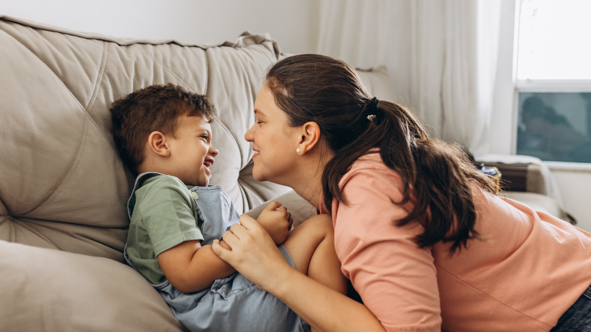 A woman and young boy playing and laughing together on a beige couch in a sunny living room.