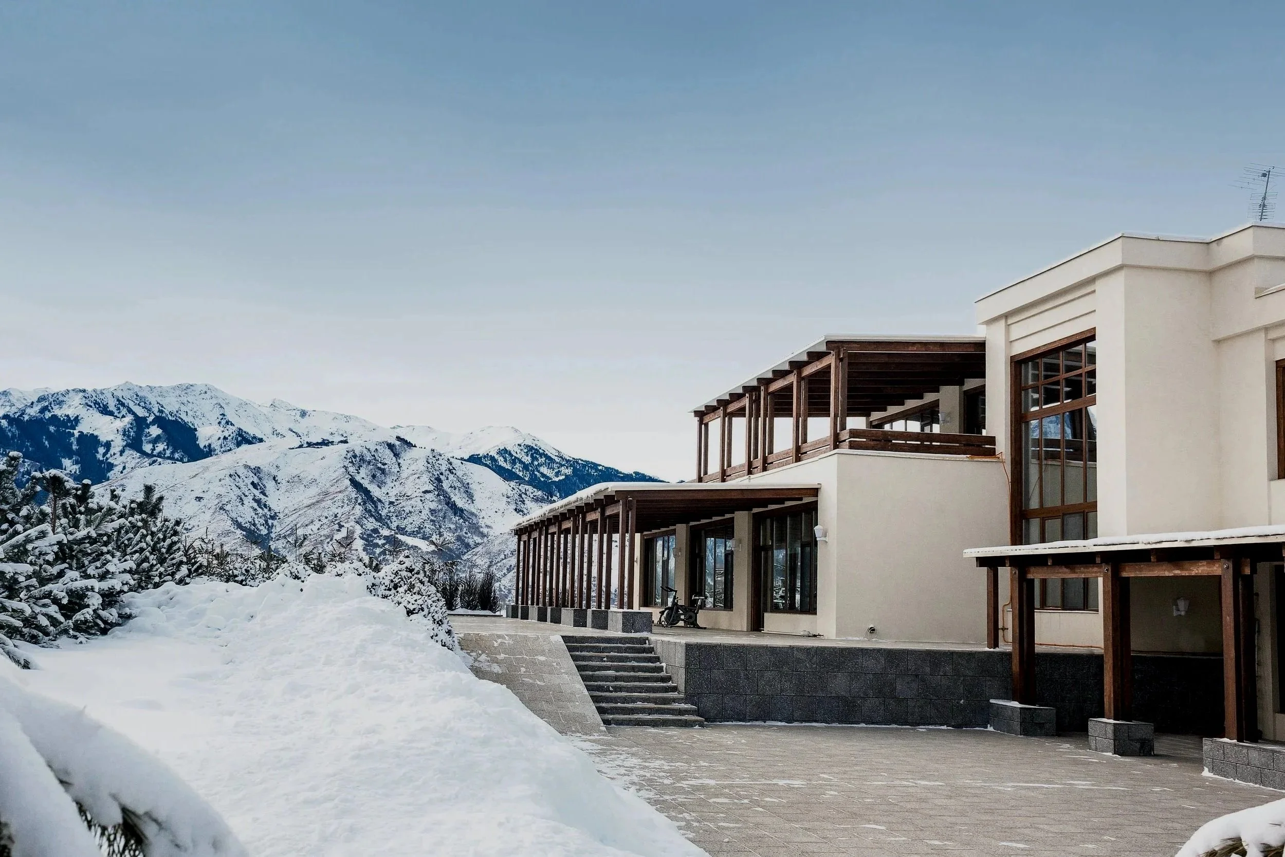 Modern house with large glass windows and wooden accents, set against snow-covered mountains and trees, with a paved driveway and snow on the ground.