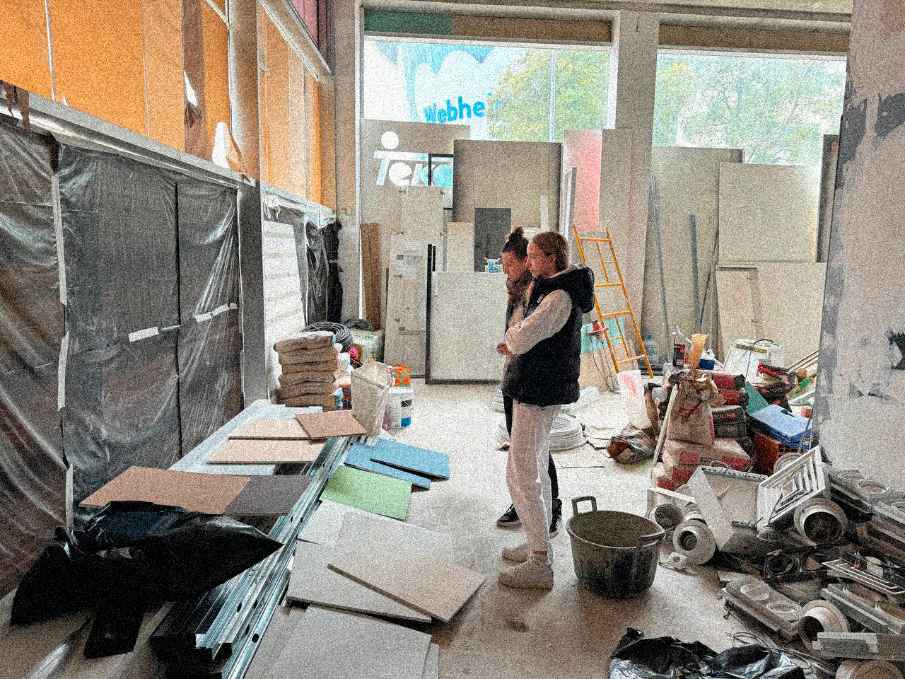 Two women are inspecting a construction site with various building supplies, tools, and materials scattered around, inside a partially finished retail space with large windows.