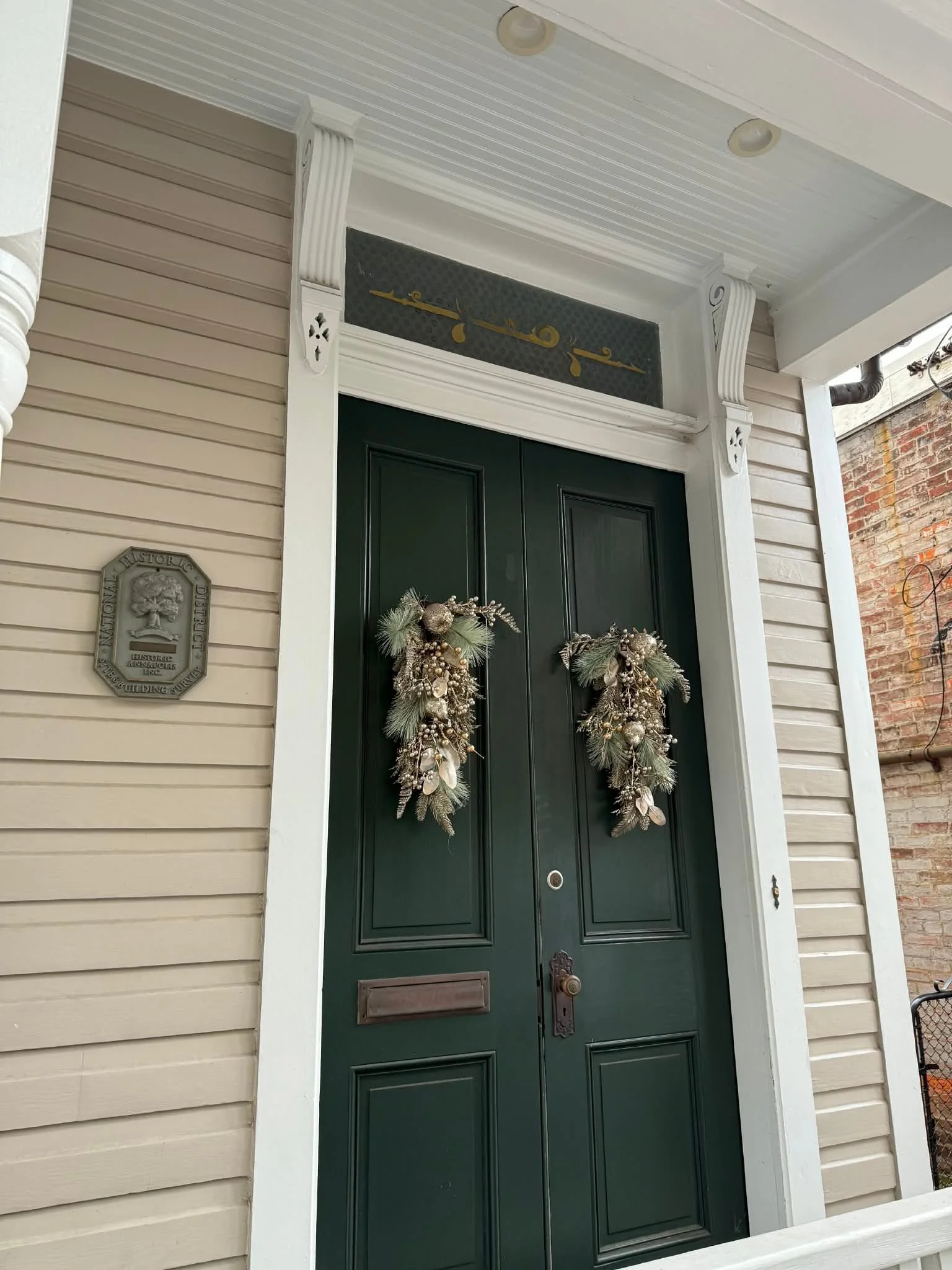 Front door decorated with two holiday wreaths on a dark green door, surrounded by beige and white siding and trim, with a plaque on the left side of the door and part of a brick wall and outdoor area visible.