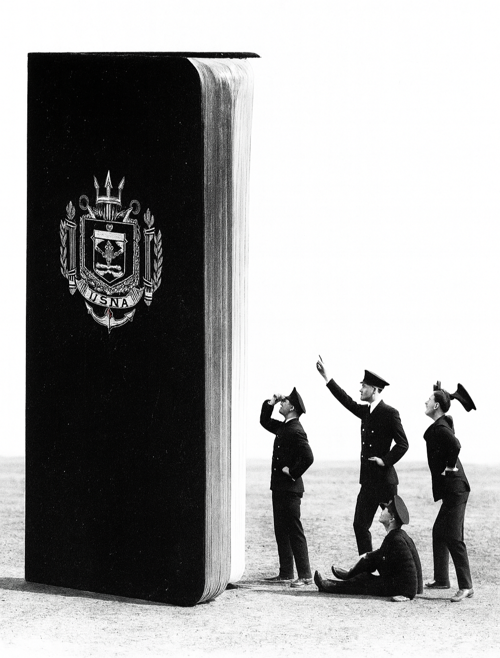 Black and white photo of five military personnel wearing dress uniforms and hats, gathered outdoors next to an extremely large book with a United States Navy emblem on the cover. One person is sitting on the ground, while the others are standing; one is pointing towards the sky.
