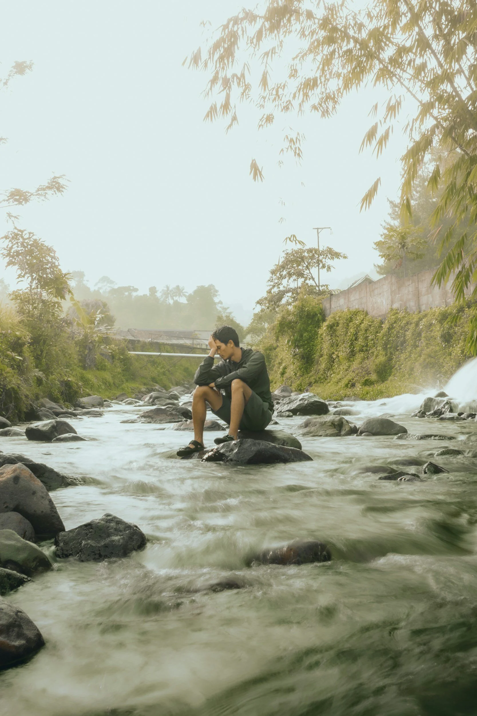 A person sitting on a rock in a river, looking down with hand on forehead, surrounded by trees and rocks, with a misty background.