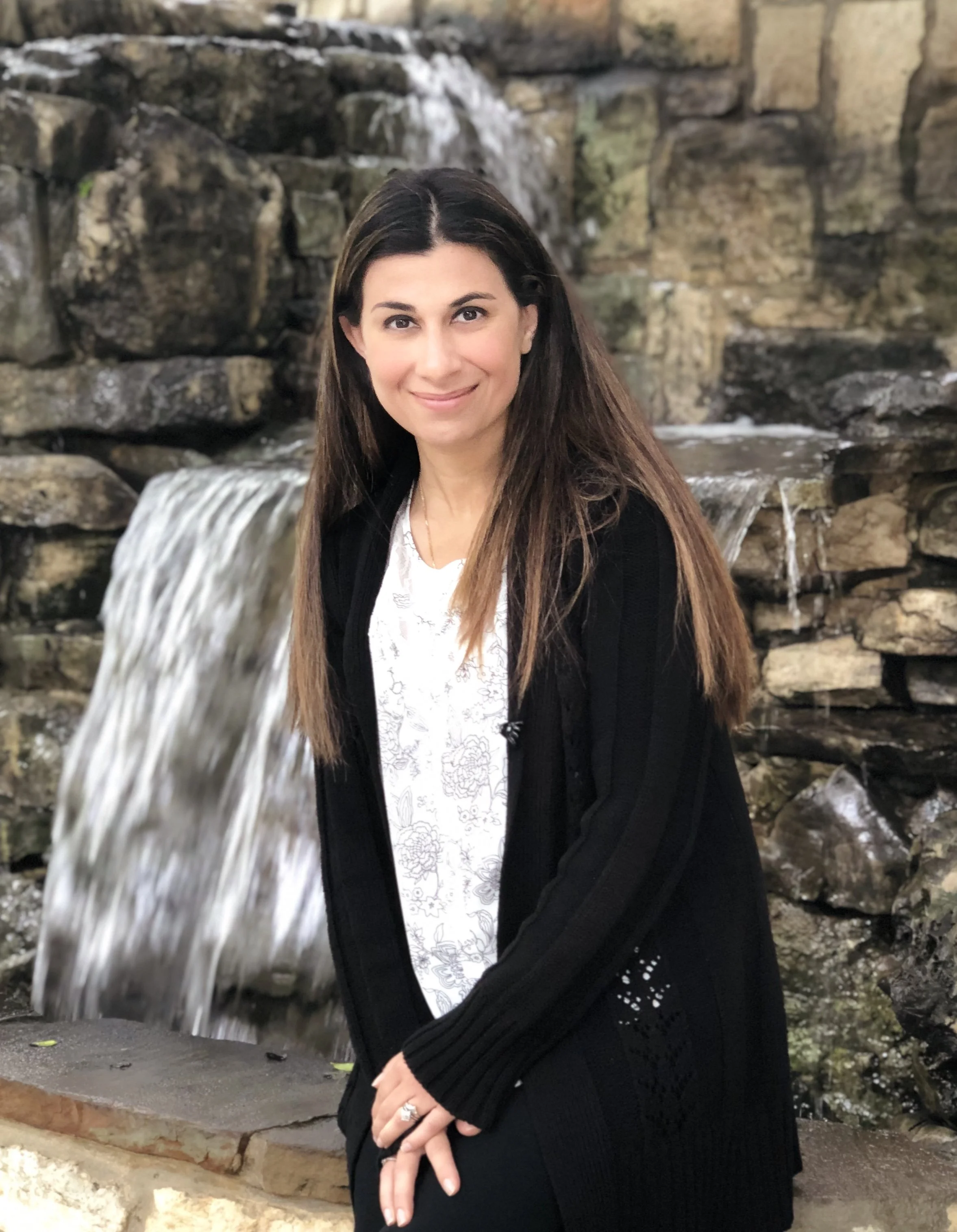 A woman with long brown hair smiling at the camera, standing in front of a small waterfall made of rocks with water flowing over them.