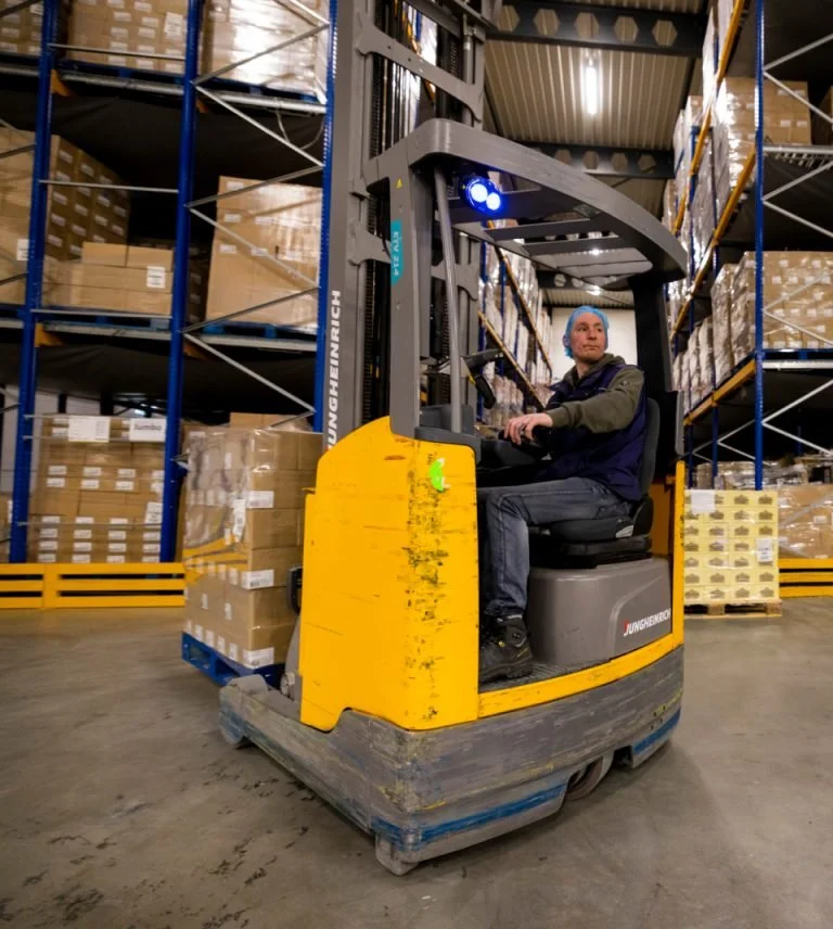 A man operating a yellow and gray electric pallet jack in a warehouse with shelves stacked with cardboard boxes.