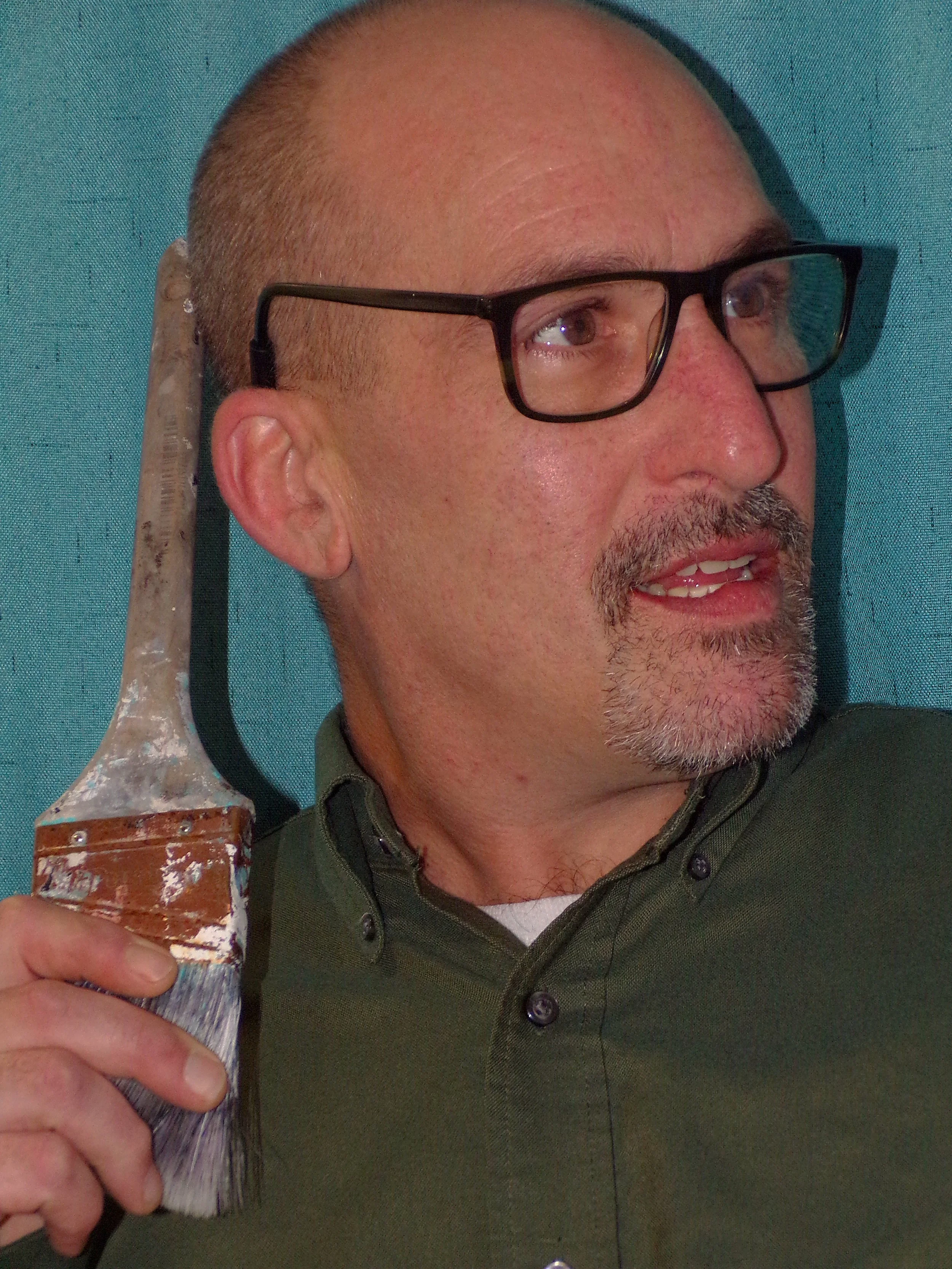 A man with glasses holding a paintbrush near his head, with a wall in the background.