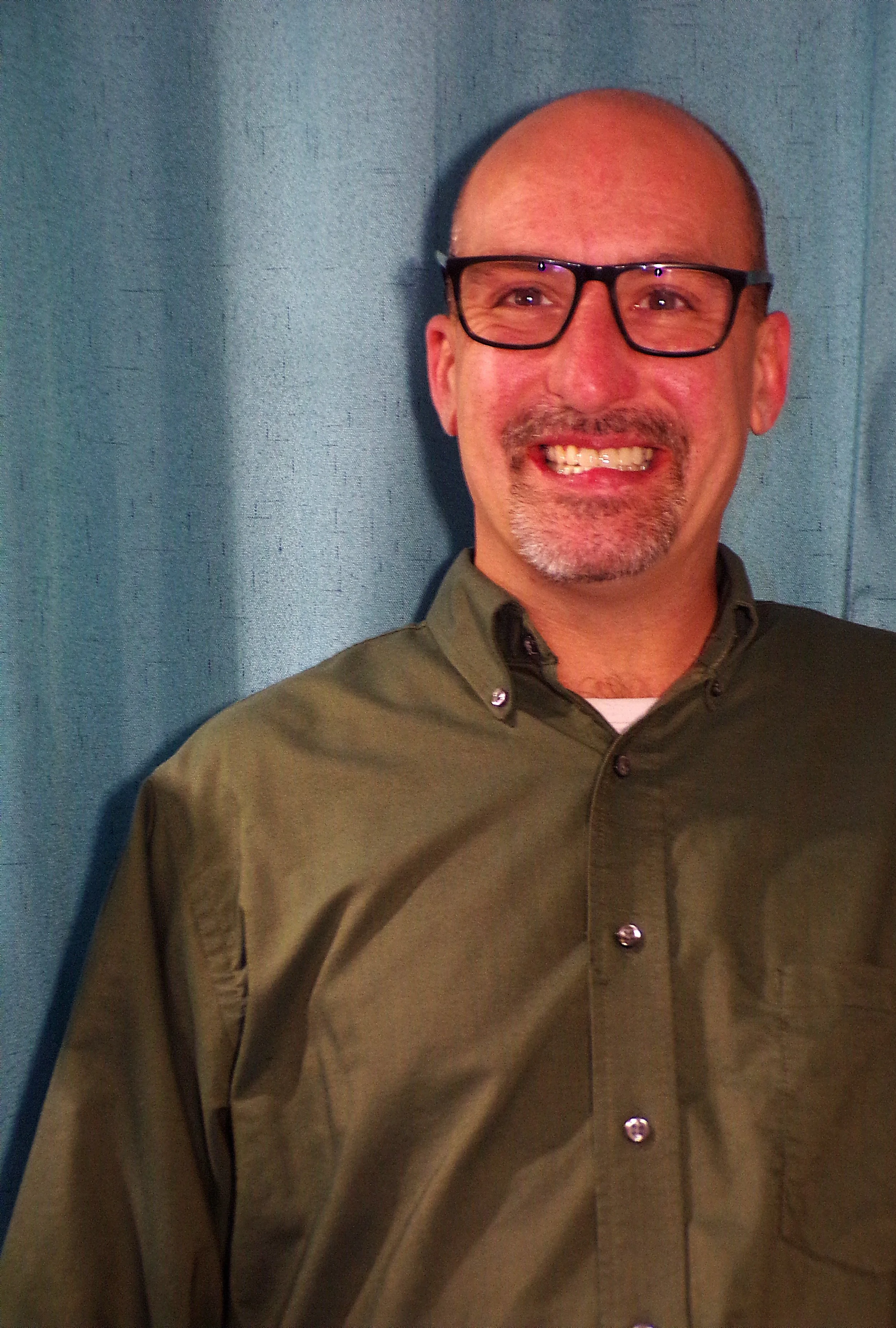 A smiling middle-aged man with glasses, a beard, and a bald head wearing a green button-up shirt, standing in front of a blue background.