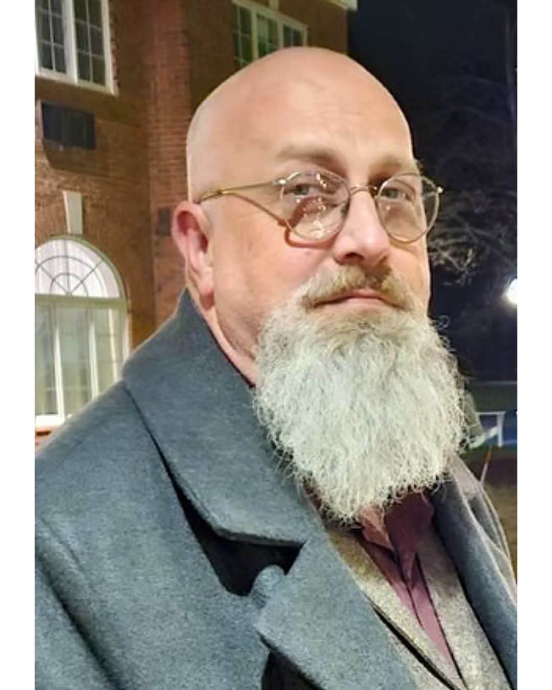 A man with a bald head, glasses, and a long white beard, wearing a gray coat, standing outdoors at night in front of a brick building with white window frames.