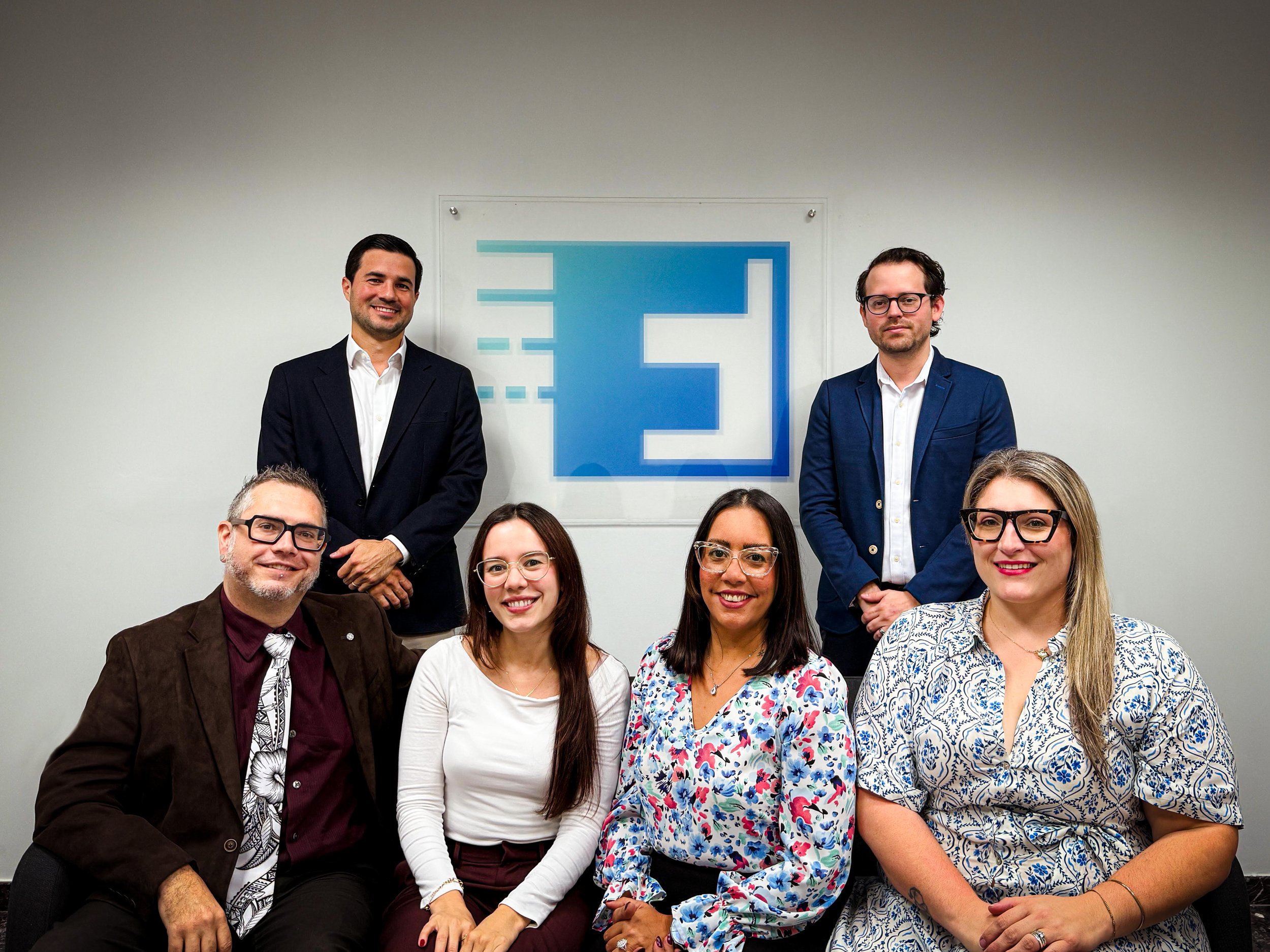 Group of seven diverse professionals, four women and three men, posing in front of a wall with a logo, some seated and some standing, dressed in business casual attire.