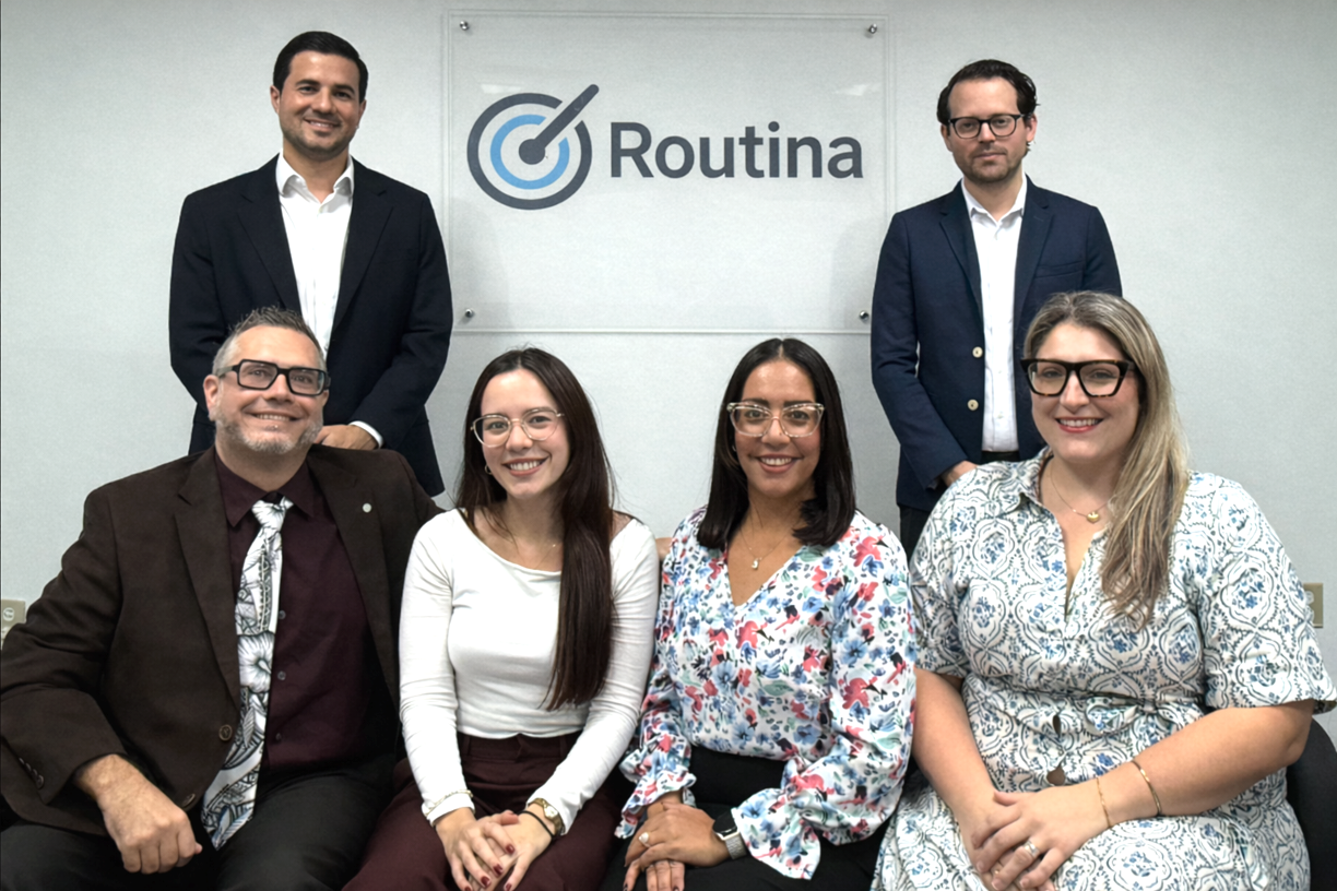 Group photo of six professionally dressed people in front of a wall with a company logo that reads 'Routina' and features a target with an arrow. Three people are seated in the front row, and three are standing behind them.