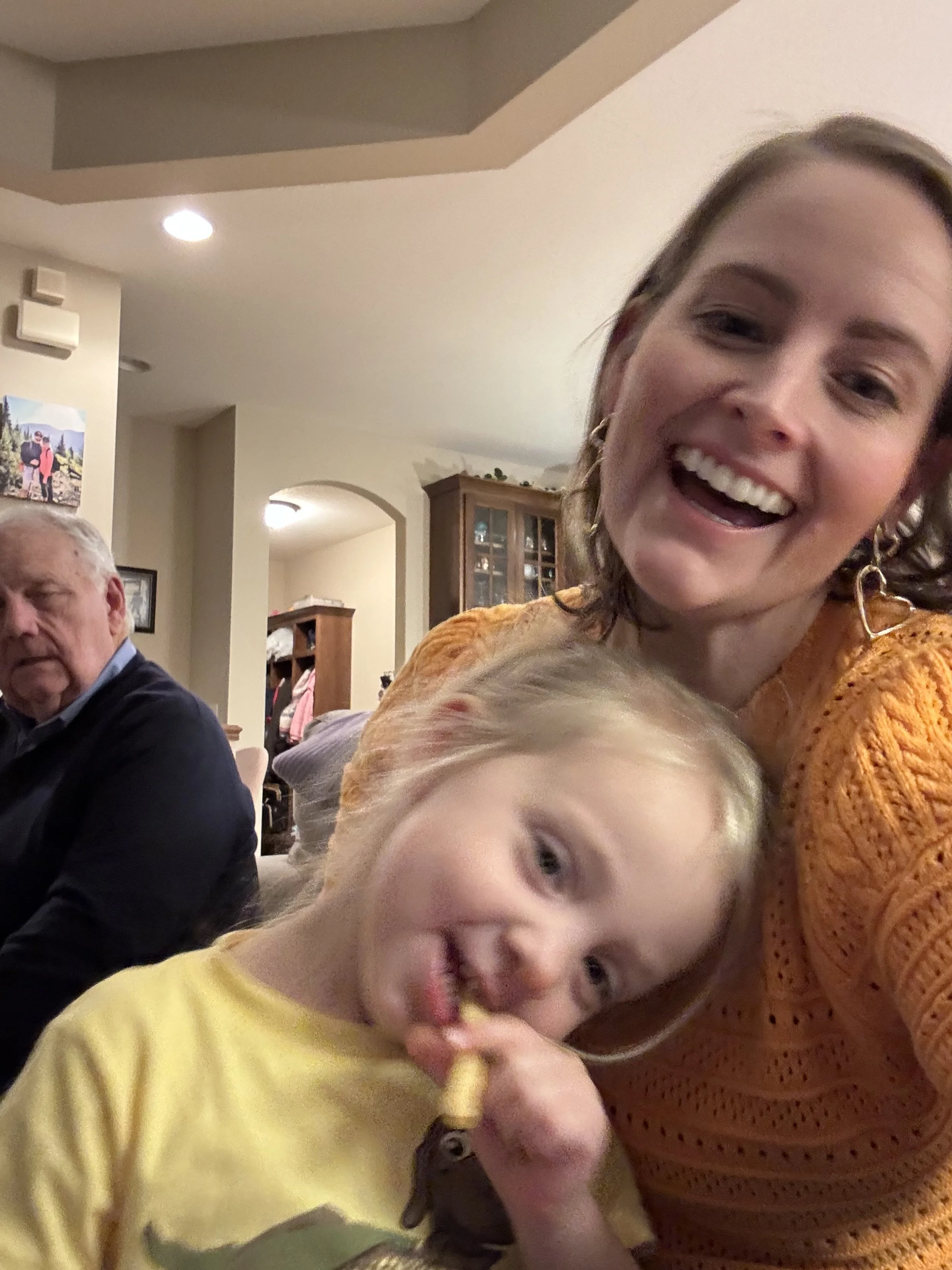 A smiling Melissa with earrings and a saffron sweater, taking a selfie with her niece Stevie in a yellow shirt, who is eating a snack. In the background, Pa G is sitting and looking away in a cozy home setting.