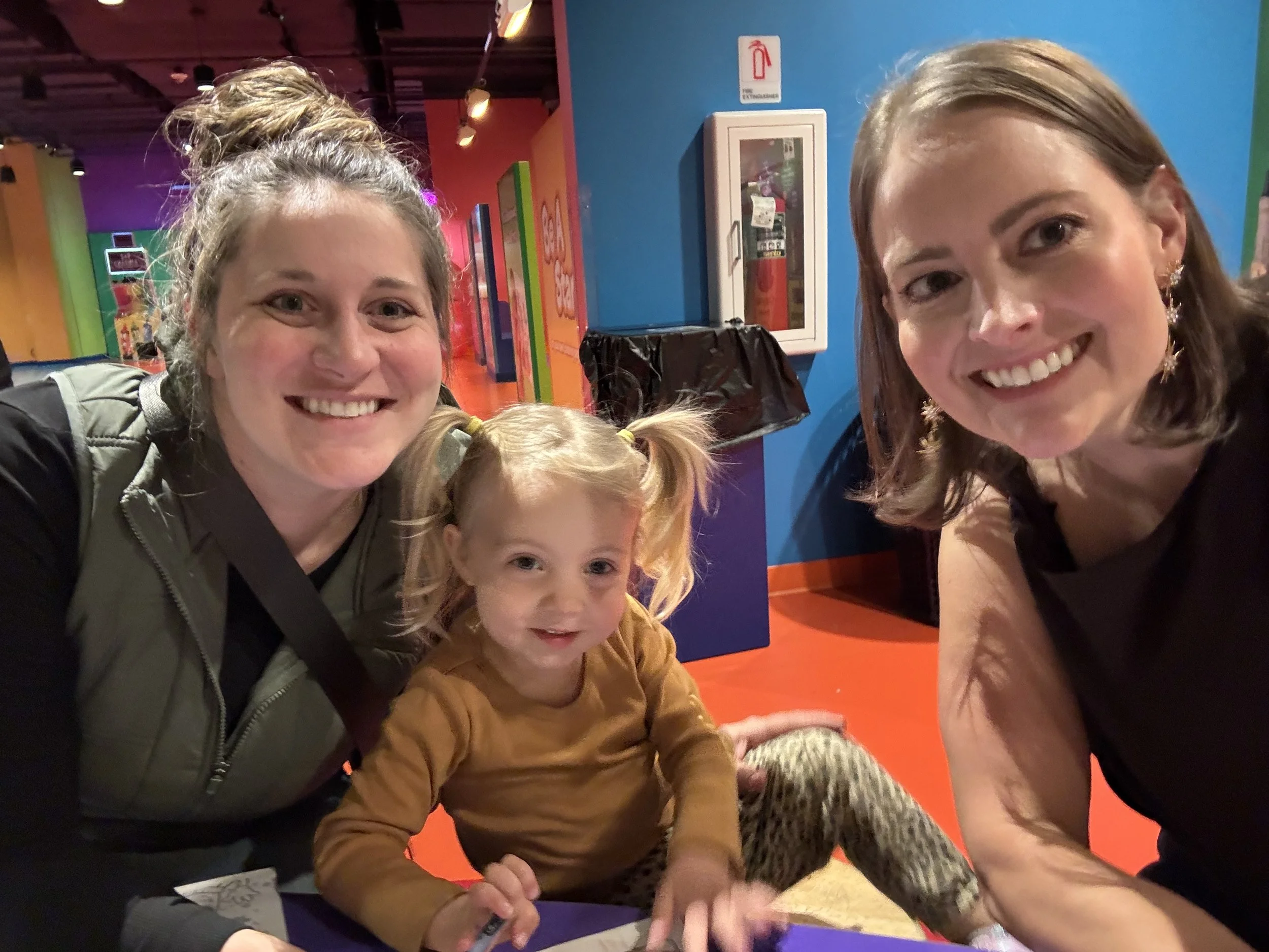 Auntie M (Melissa) and Mom, Callie, with toddler Stevie, are taking a selfie inside the Crayola Experience at The Mall of America in Minneapolis, Minnesota. All are smiling and enjoying the outing.