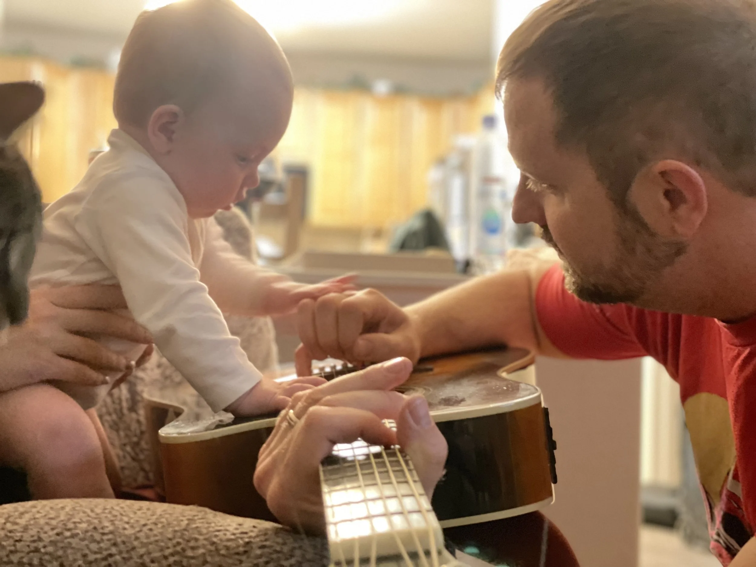 Matt Moss and his baby daughter Stevie are sharing a moment where Matt is helping the baby hold an acoustic guitar. Matt is focused on the guitar while baby Stevie is also reaching for it in a cozy, home setting.