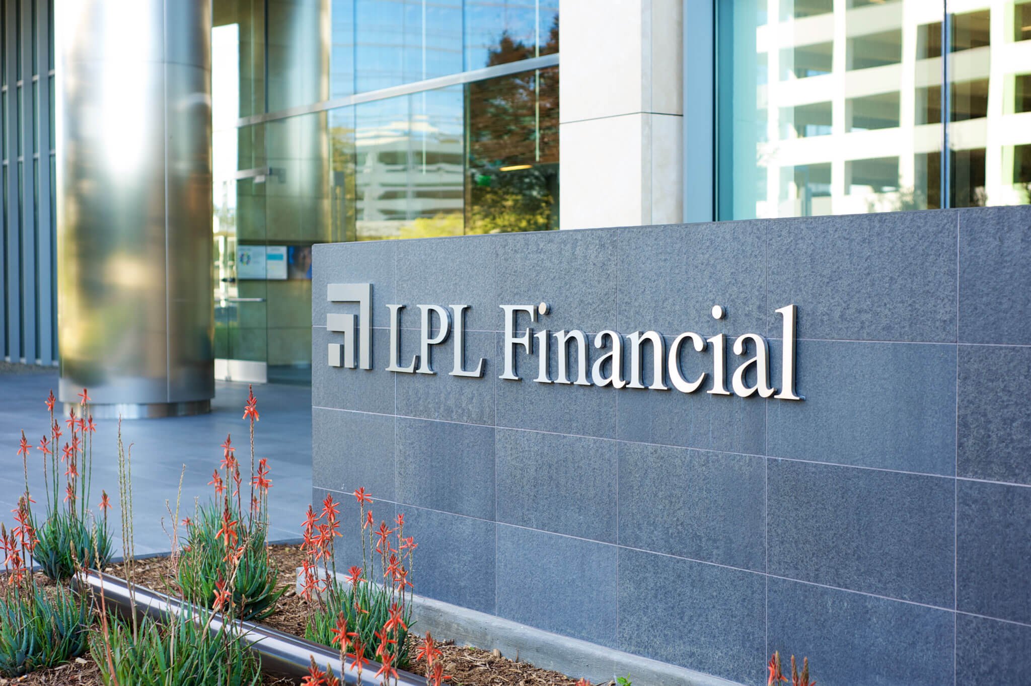 Exterior view of LPL Financial building with a modern design, gray stone wall, and silver signage, front garden with red flowers, glass windows reflecting trees, and a cylindrical metallic column. San Diego, California.