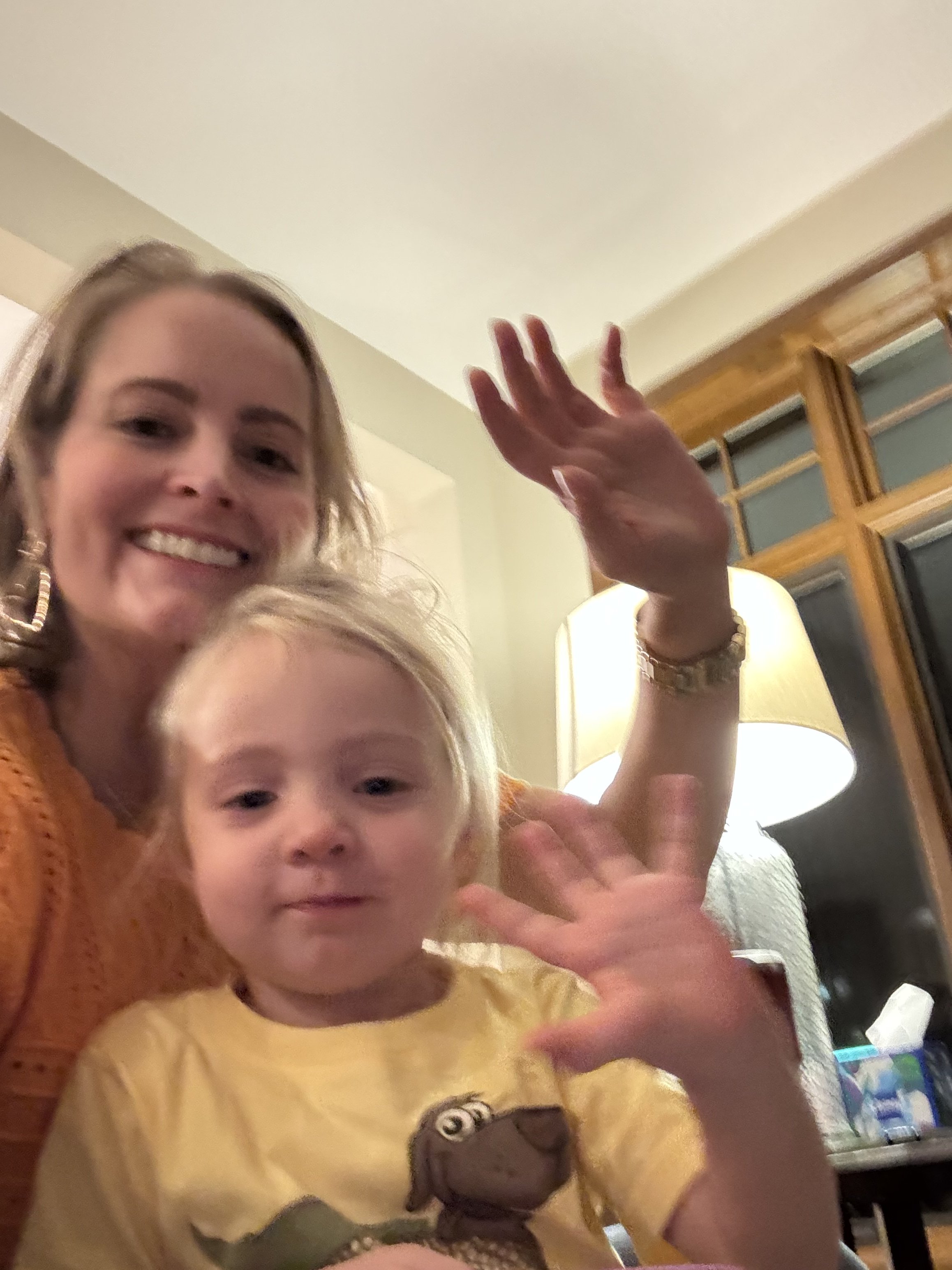 A smiling woman with blonde hair and earrings waving her hand, standing near a young child with blonde hair, both indoors with warm lighting and a window in the background.