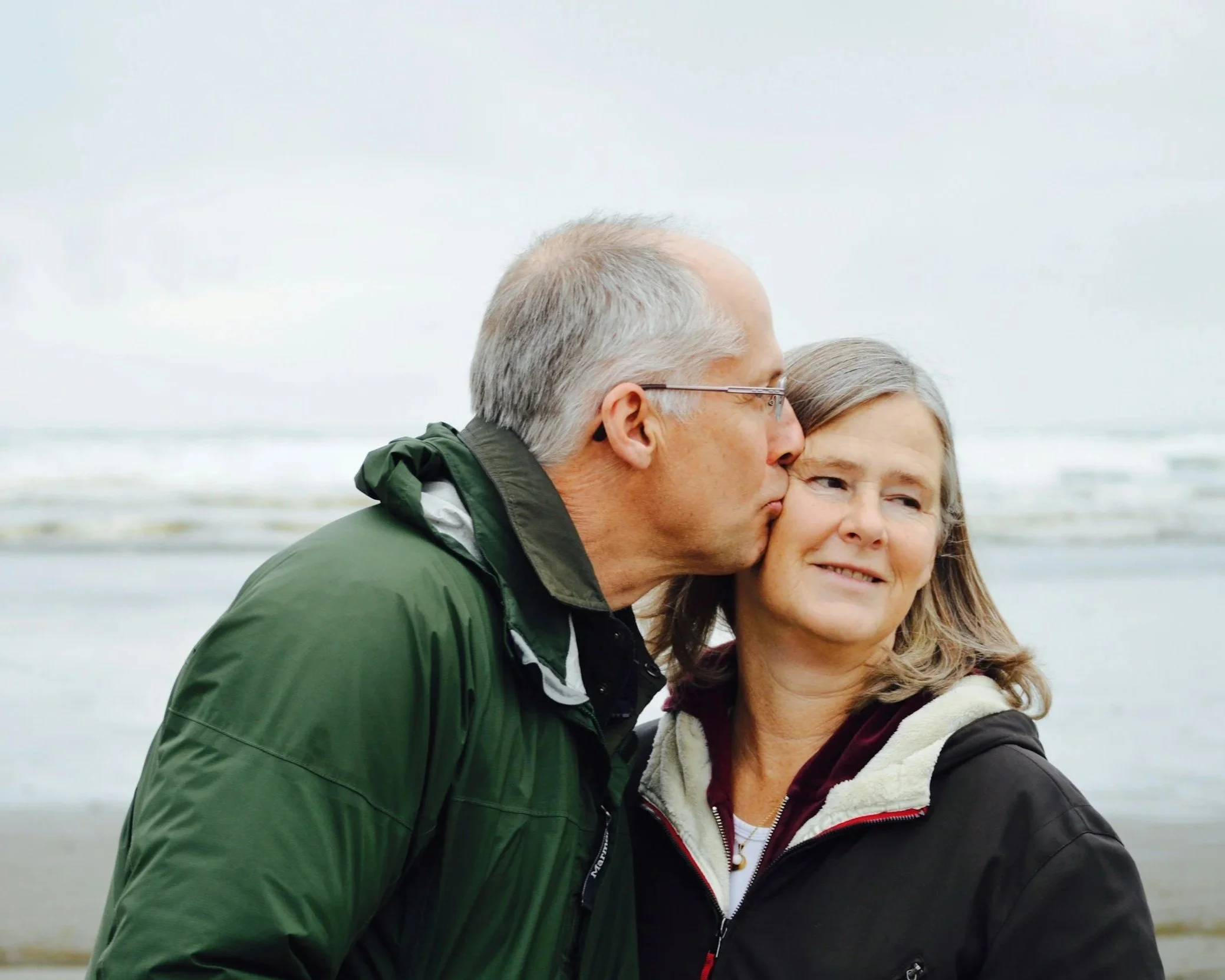 An elderly man with gray hair and glasses kisses a woman on the cheek at the beach. The woman has shoulder-length gray hair and is smiling softly.