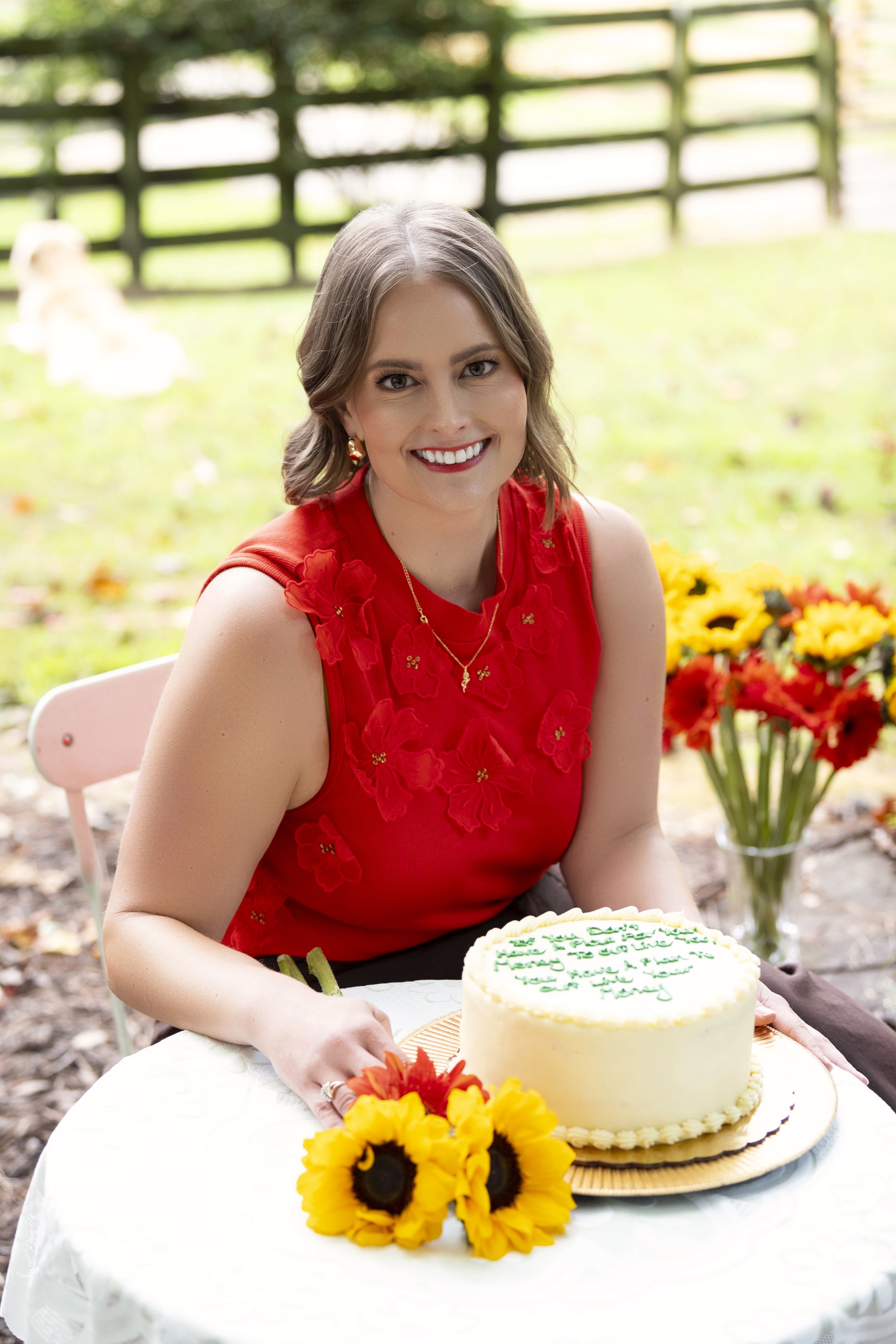 A woman with shoulder-length brown hair, wearing a red sleeveless top with floral embroidery, sitting at a white table outdoors. She is smiling and posing with a white frosted birthday cake with green writing, a bouquet of sunflowers, and a background with a wooden fence and colorful flowers.
