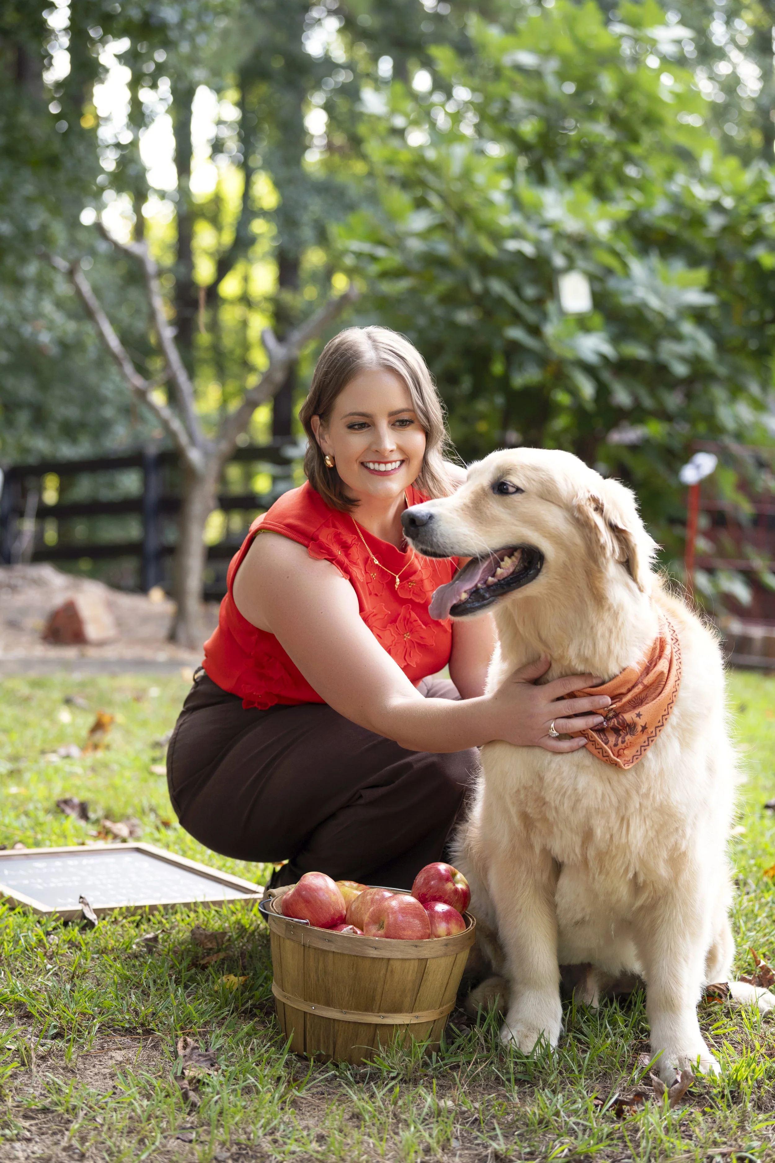 Melissa is crouching on the grass, smiling, with Riley, the golden retriever, wearing an orange bandana. There is a basket of apples and a chalkboard on the ground nearby, with trees and a wooden fence in the background.