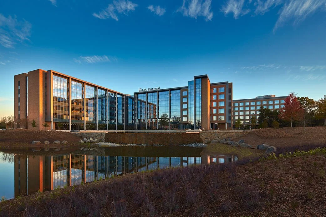 A modern office building with a glass facade, reflecting in a small pond in the foreground, under a clear blue sky.