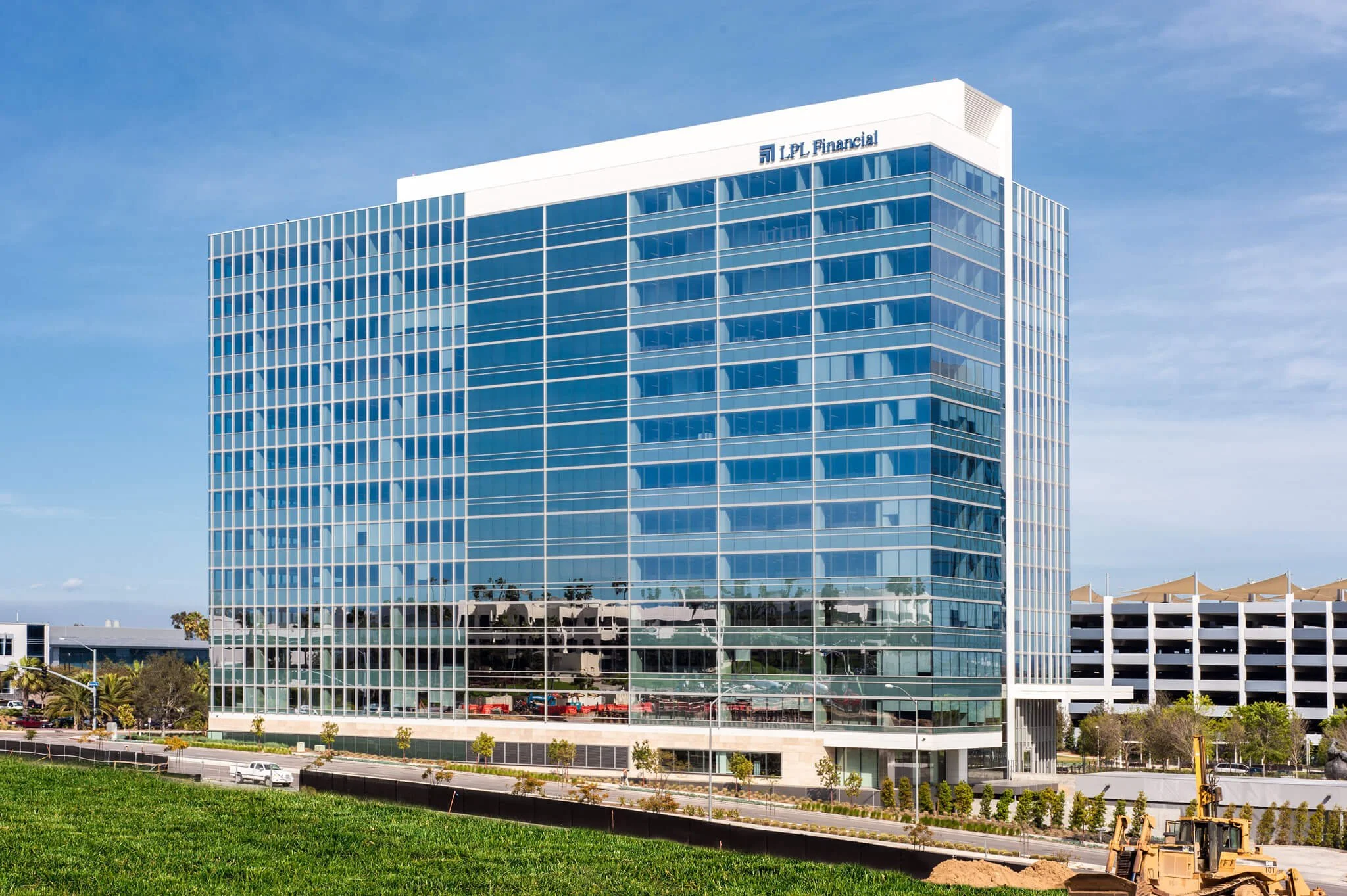 A modern multi-story office building with glass windows and the logo "LPL Financial" on the top corner, under a partly cloudy blue sky. There is a parking structure on the right and some greenery in the foreground.