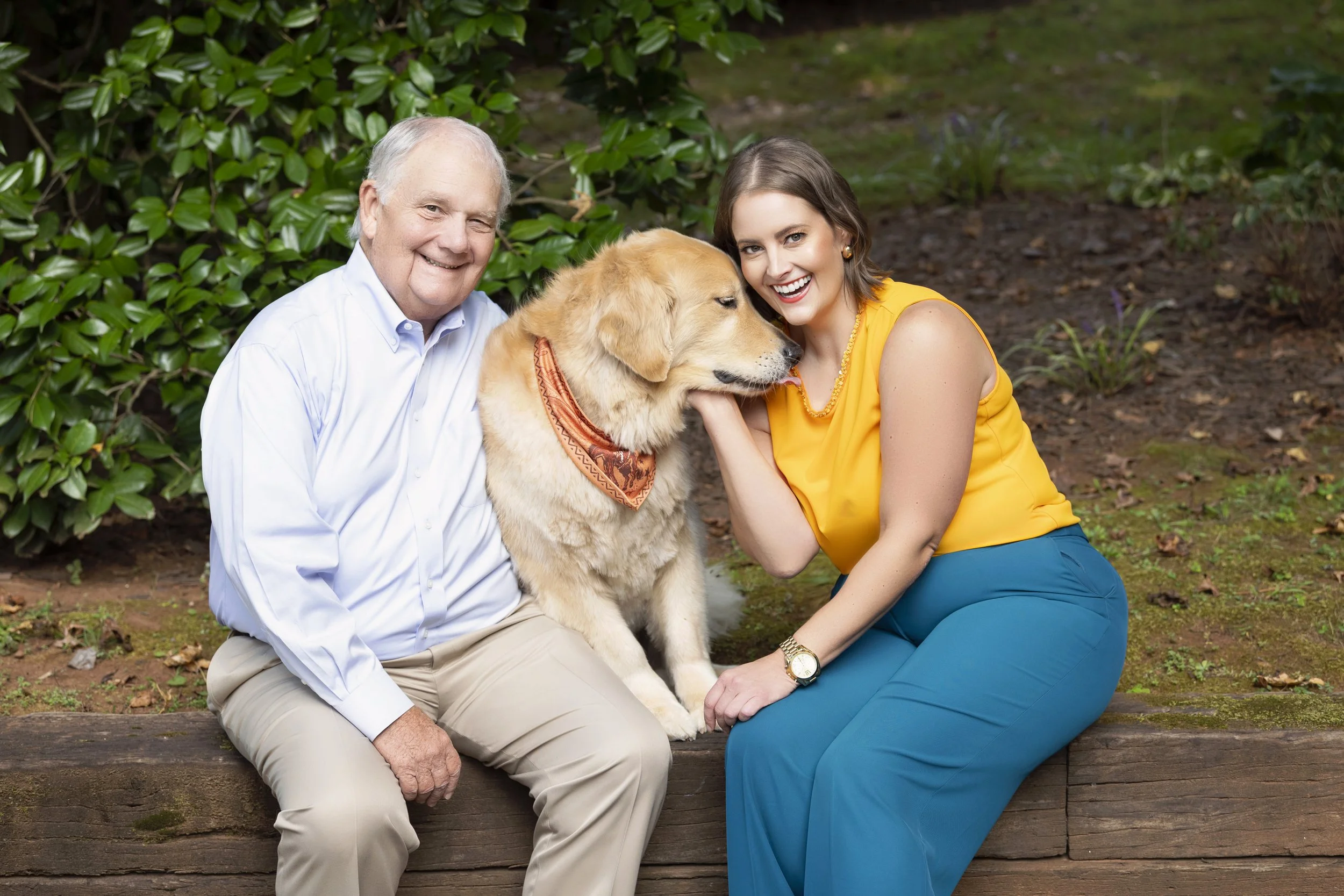 Grady Moss, Melissa Moss, and golden retriever Riley are sitting outside. Grady is wearing a white shirt and beige pants, Melissa is wearing a saffron top and kingfisher blue pants, and Riley has an orange bandana. They are smiling and happy.