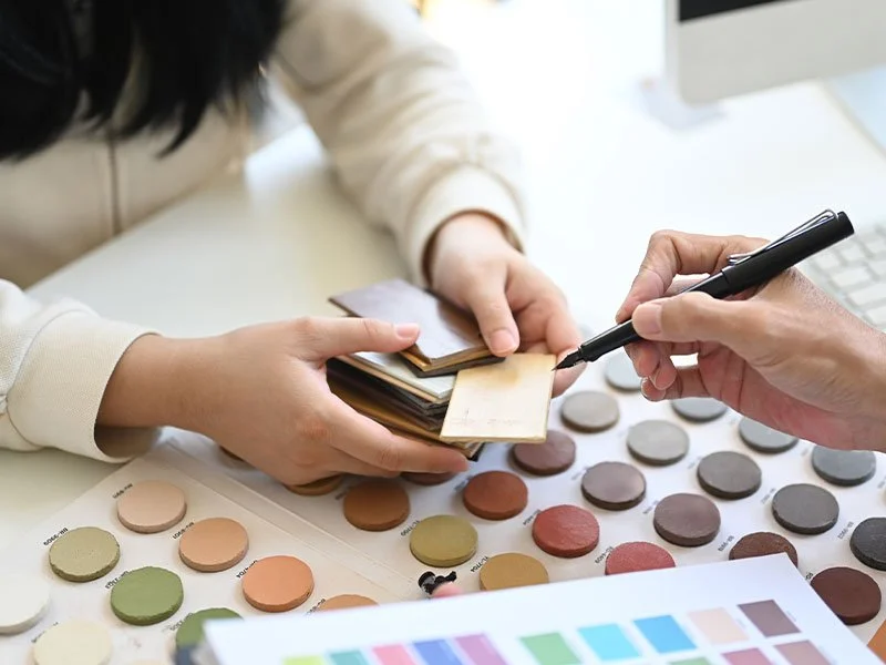 Two people are discussing fabric color samples, with one person holding small fabric swatches and the other writing on a fabric sample, surrounded by color palettes and design materials.