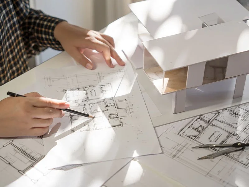 Person working on architectural plans with a model house and drafting tools on a table.