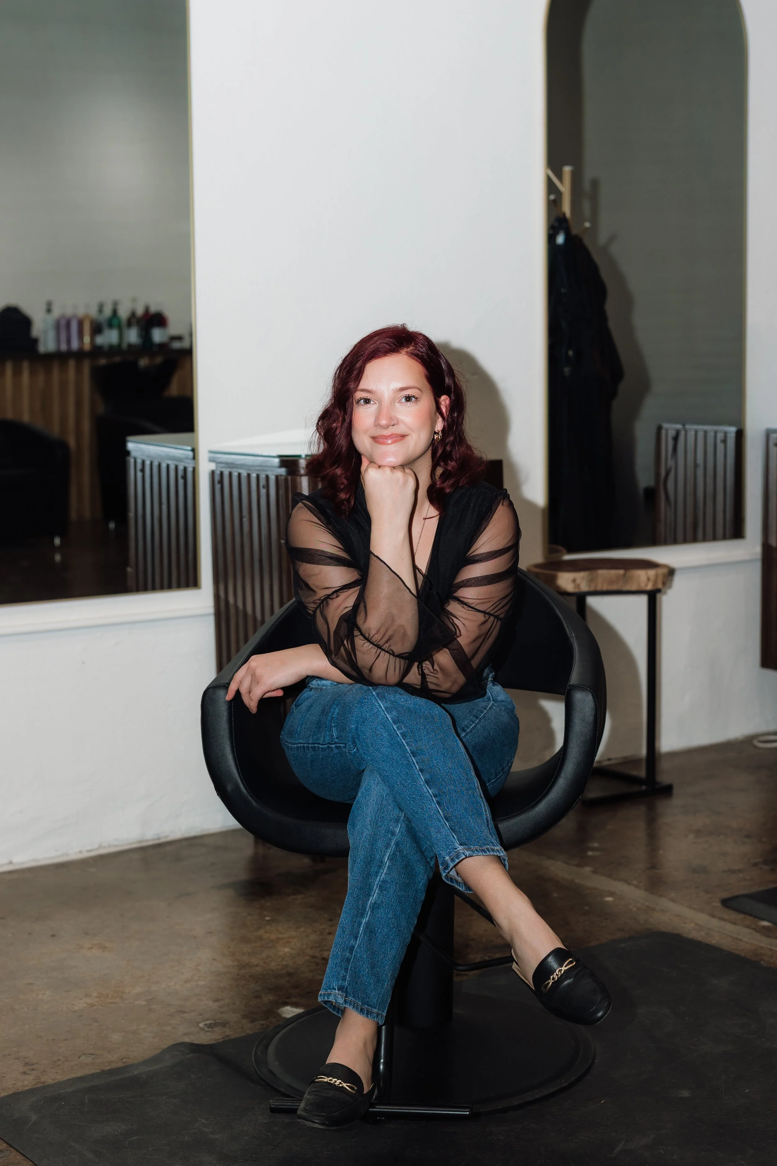A young woman with a cheerful smile, wearing a tan headband, gold hoop earrings, a necklace with a snake-shaped pendant, and a black top with sheer floral sleeves, standing against a white brick wall.