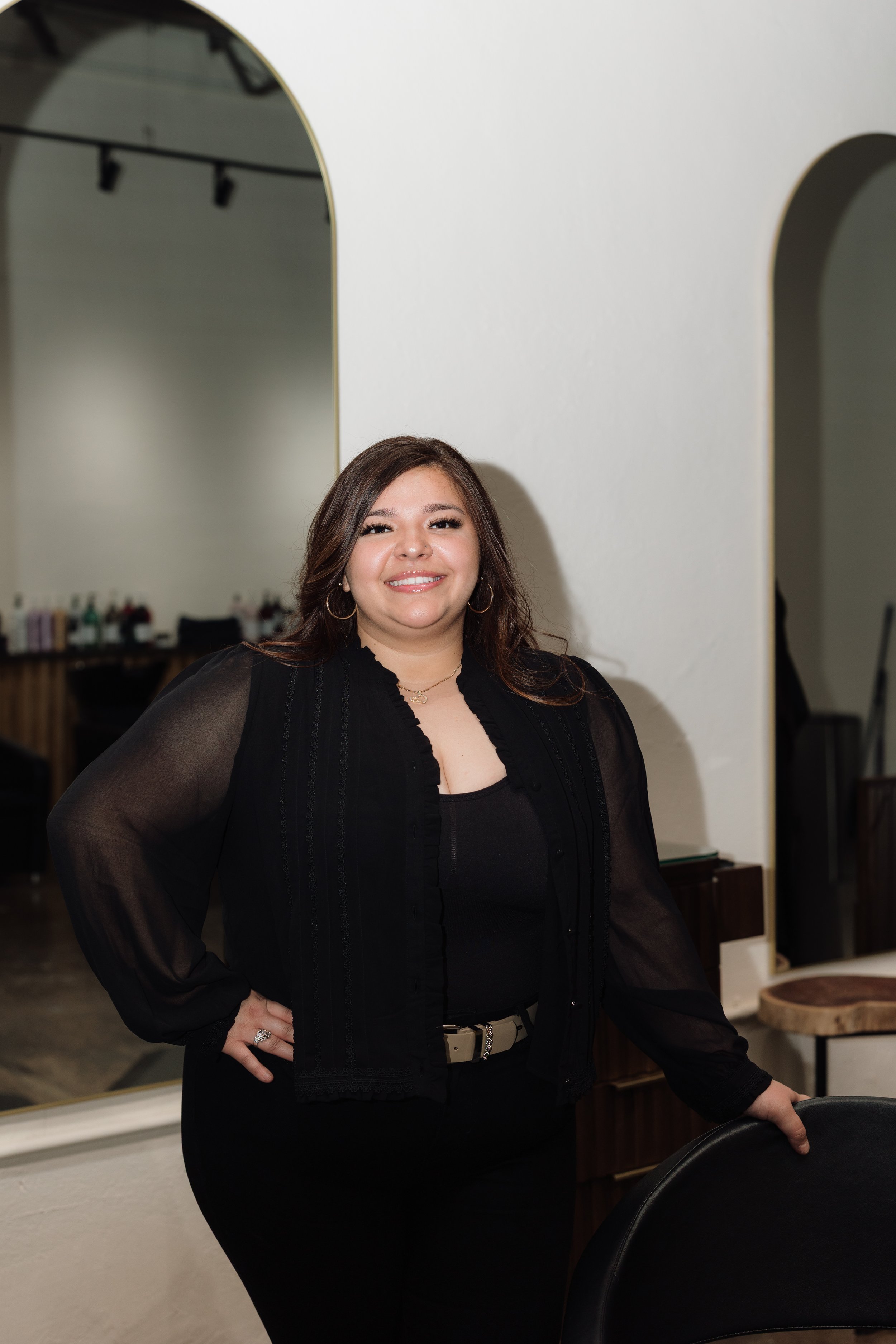A woman with dark, wavy hair smiling in front of a white brick wall.