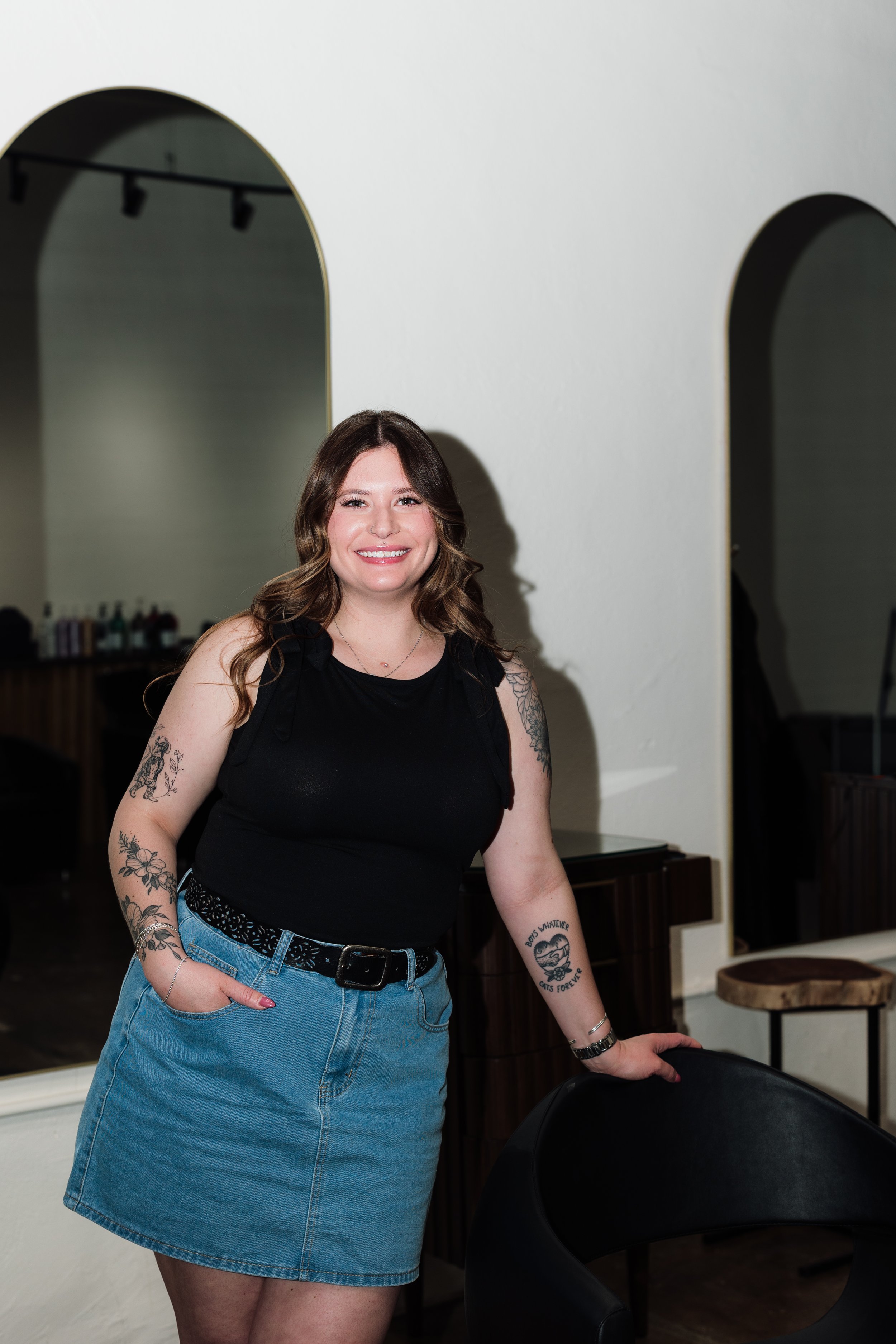 A young woman with long brown hair and a nose piercing smiling at the camera, standing in front of a white brick wall, wearing a black sleeveless top and jewelry, with a floral tattoo on her right shoulder.