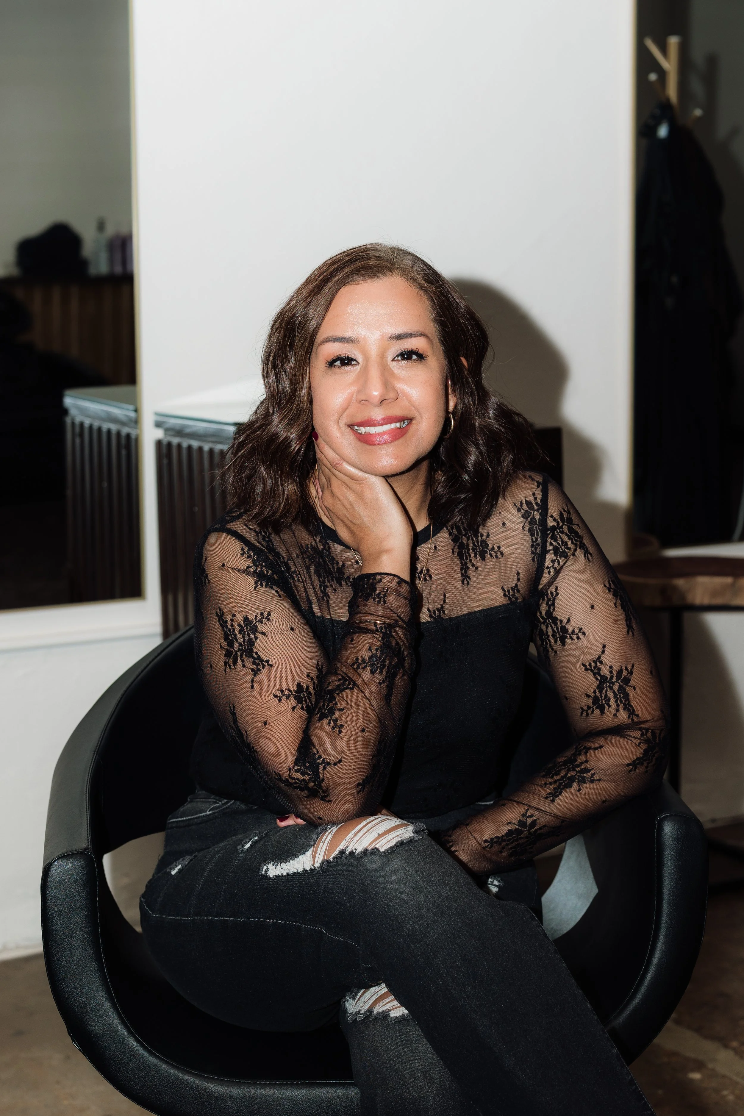 A woman with shoulder-length wavy brown hair smiling and wearing a dark blue top, standing in front of a white brick wall.