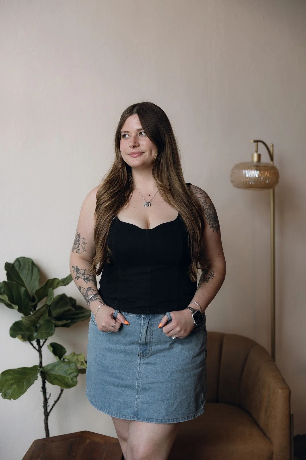 A young woman with long brown hair and a nose piercing smiling at the camera, standing in front of a white brick wall, wearing a black sleeveless top and jewelry, with a floral tattoo on her right shoulder.