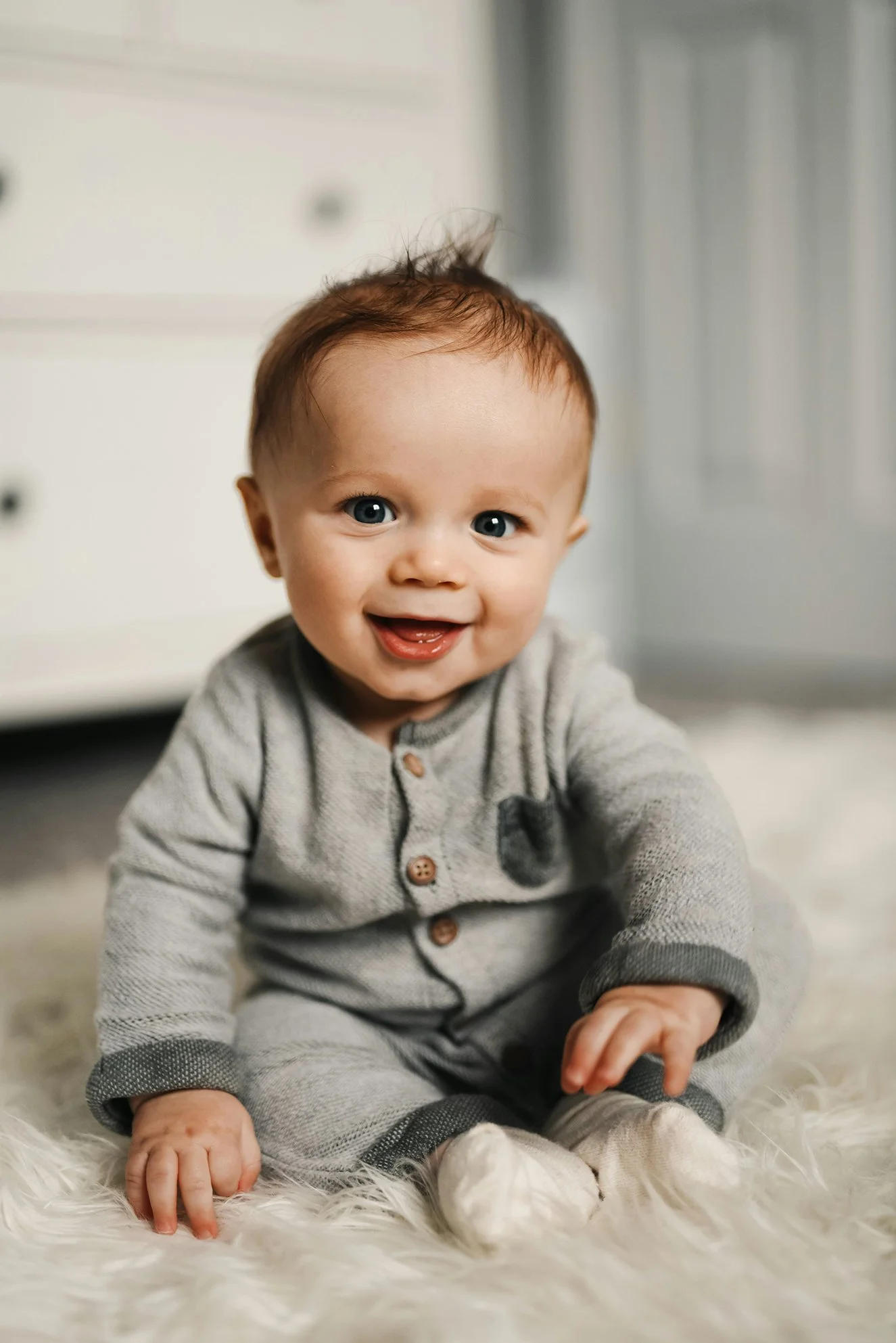 A smiling baby with brown hair and blue eyes, wearing a gray onesie with brown buttons, sitting on a soft, white fur rug.