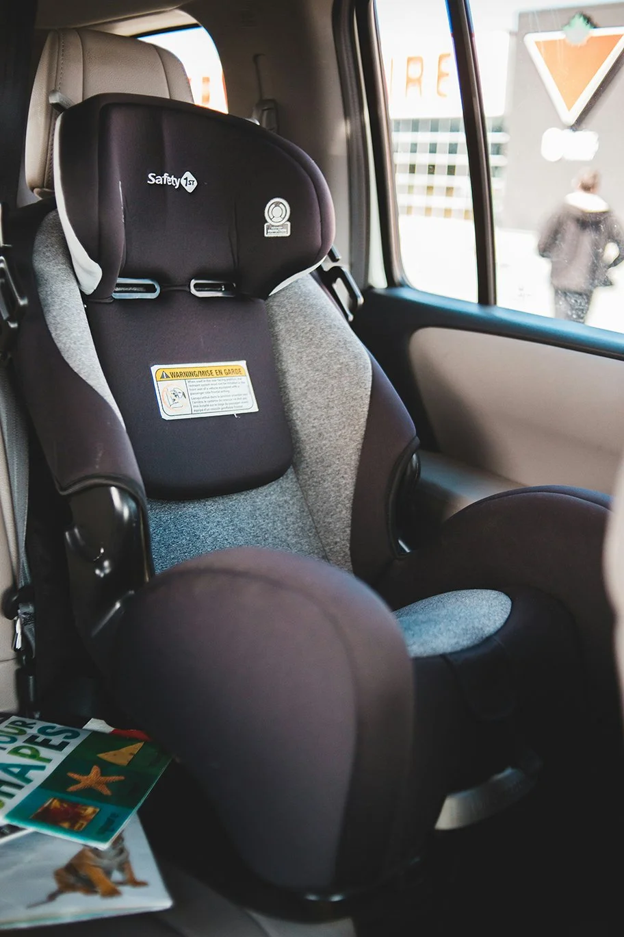 Child car seat installed in the back seat of a vehicle next to a window, with a person walking outside visible through the window.