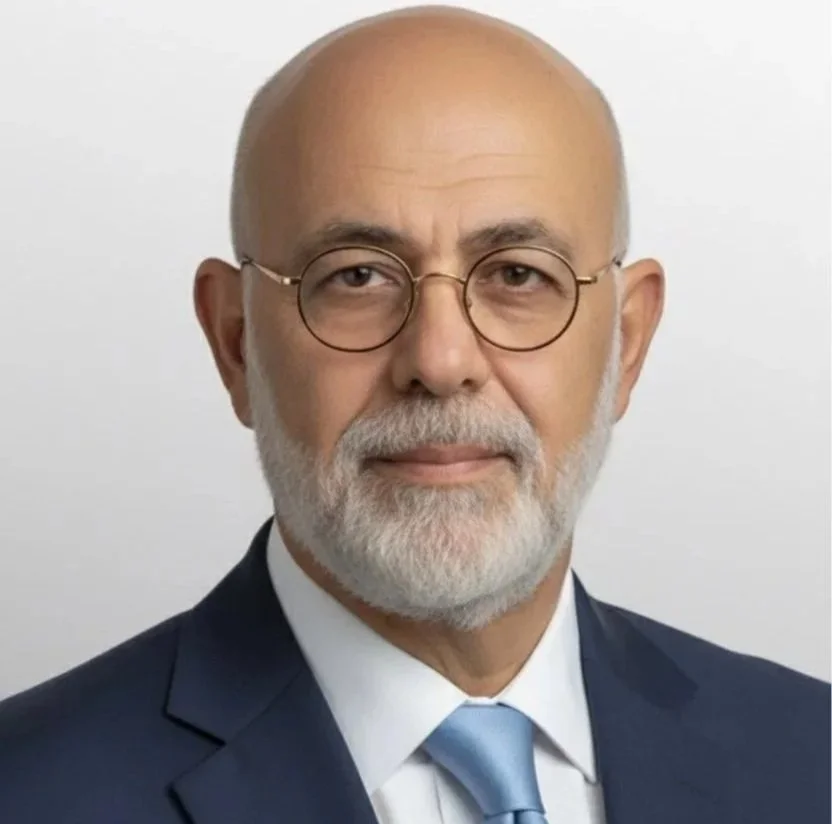 A middle-aged man with a bald head, glasses, and a white beard, wearing a suit and tie, against a plain light background.