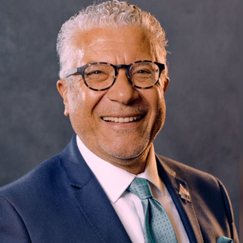Portrait of an older man with gray curly hair, wearing glasses, a navy blue suit, white shirt, and light blue tie, smiling against a gray background.