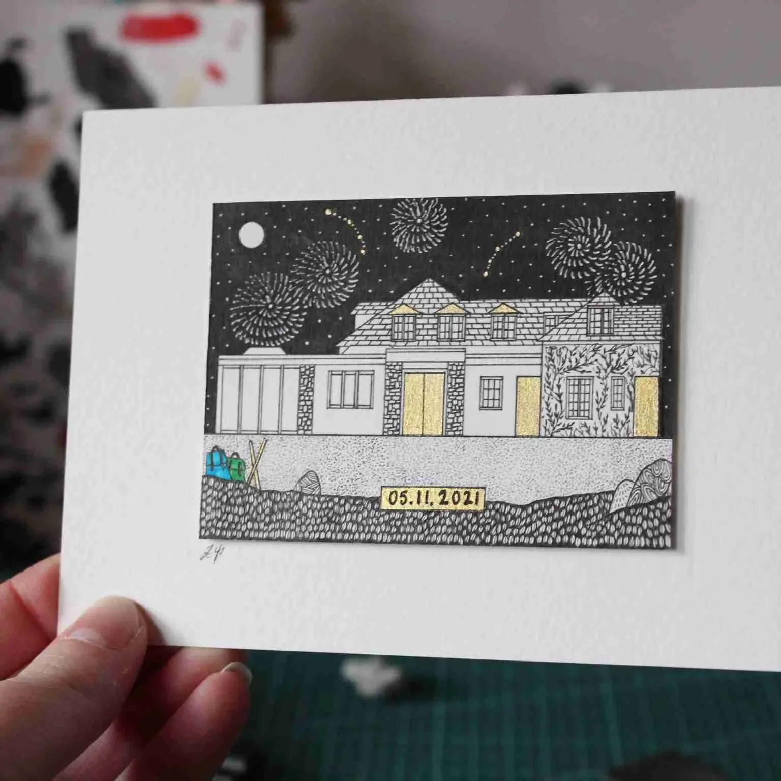 Illustration of a house with fireworks in the night sky, a date '05.11.2021' on a yellow background at the bottom, and a hand holding the card.