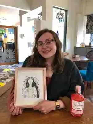 A woman with glasses and long brown hair sitting at a table, smiling and holding a framed picture of a dog. There is a red bottle on the table.