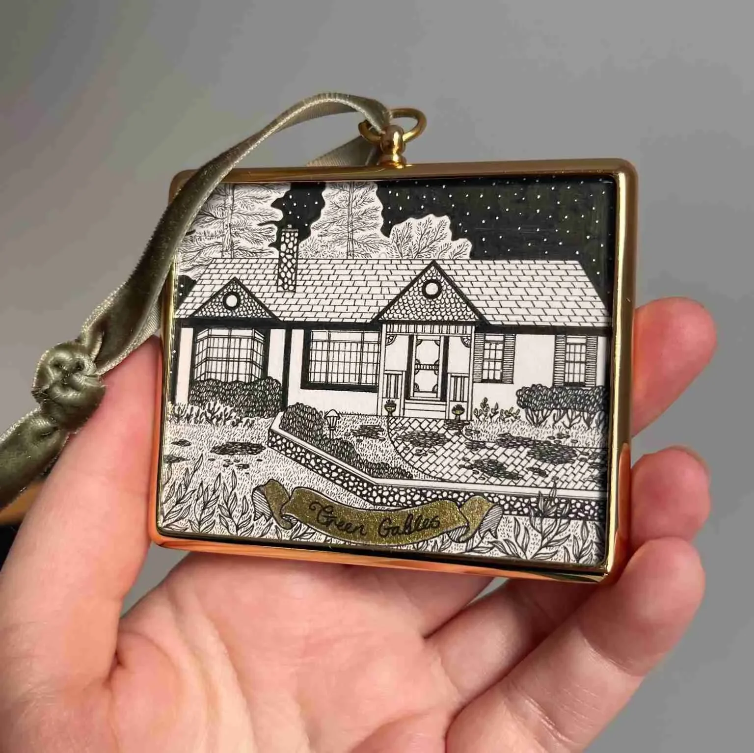 A decorative compact mirror with a black and white illustration of a house and garden on the lid, with a gold border and a ribbon with the words 'Seven Gables'.