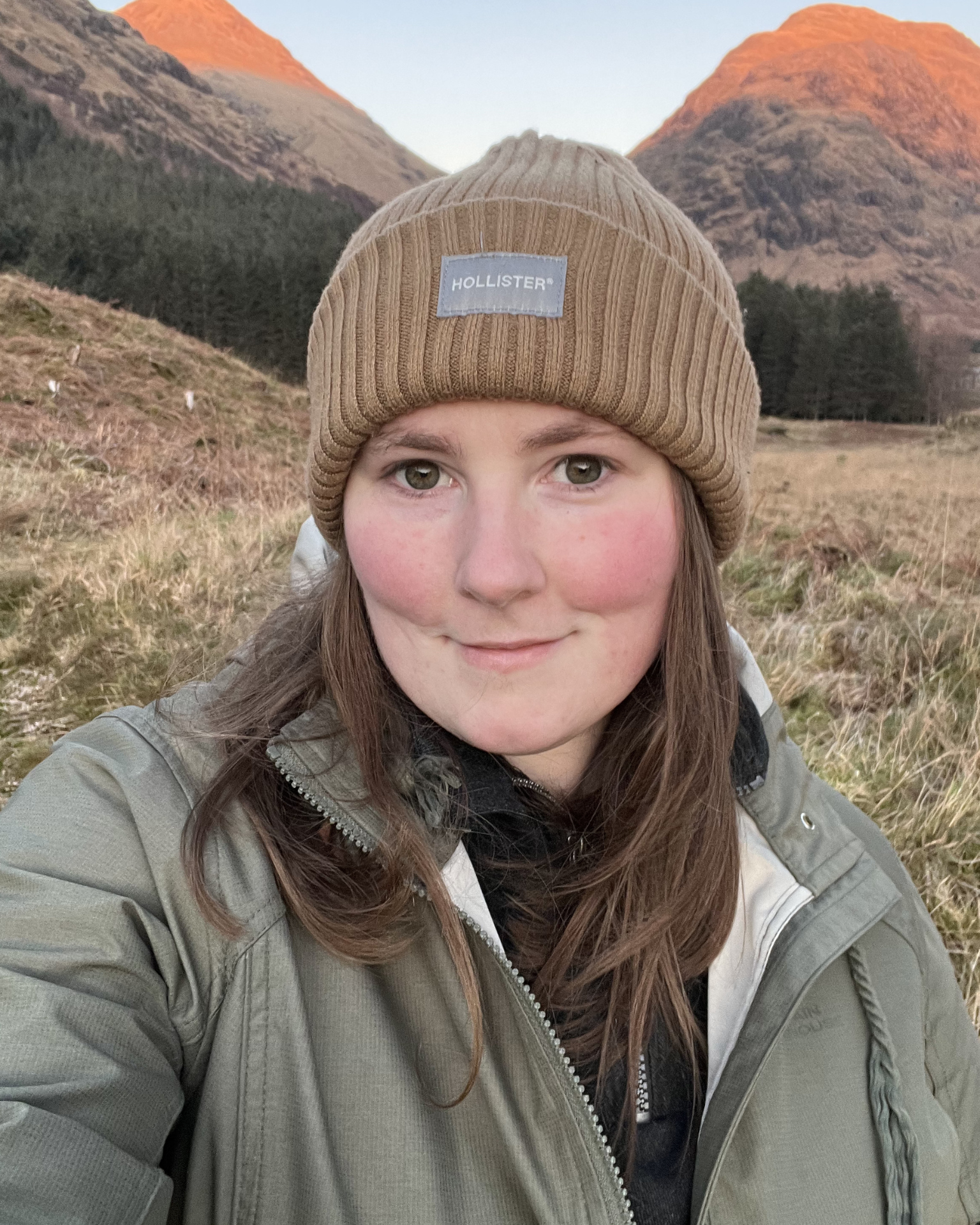 A woman with light brown hair, wearing a tan Hollister beanie and a gray jacket, taking a selfie outdoors with a mountainous landscape at sunset in the background.