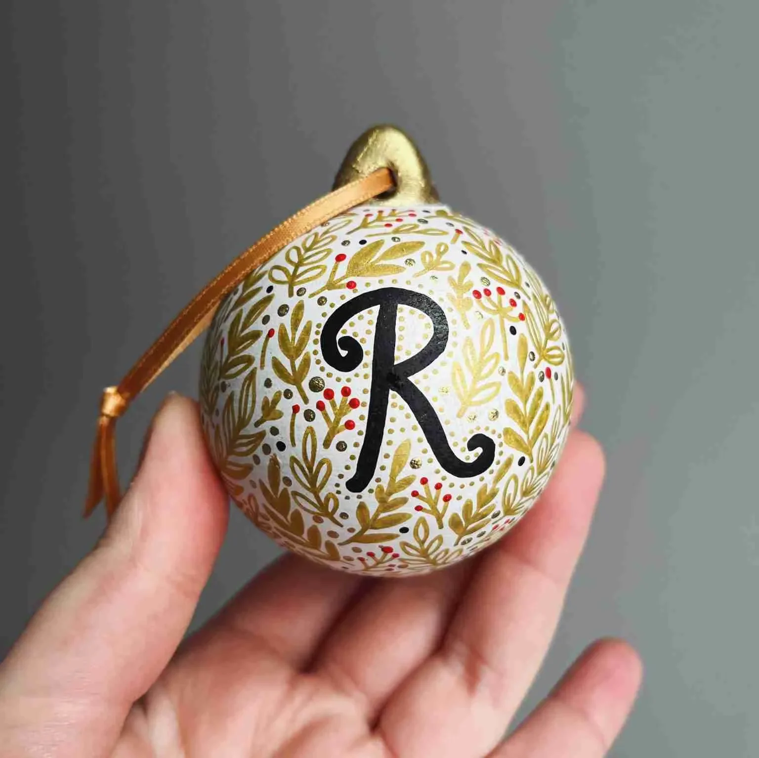 Hand holding a decorated Christmas ornament with a large black letter 'R' on a white background, surrounded by gold floral patterns, red berries, and gold accents, with a gold ribbon loop at the top.