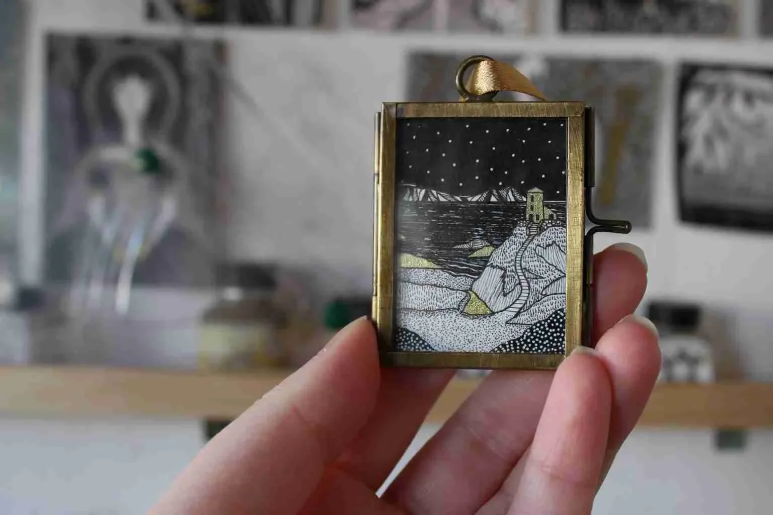 Hand holding a small, rectangular, gold-colored picture frame with an illustrated black and white landscape scene featuring a house along a rocky coastline and a night sky with stars.
