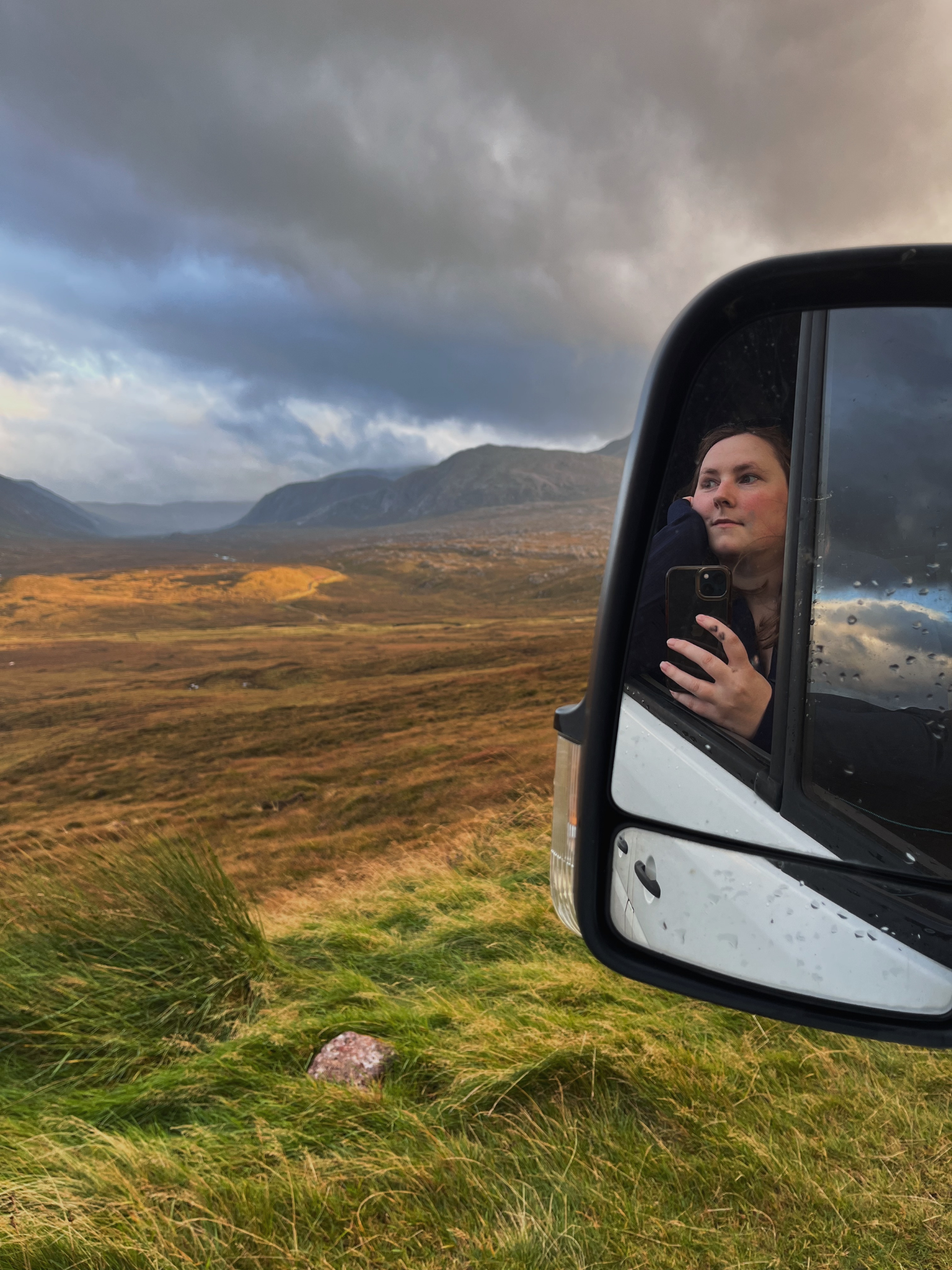 A young woman taking a selfie in a vehicle's side mirror, with a landscape of rolling hills and mountains under a cloudy sky in the background.