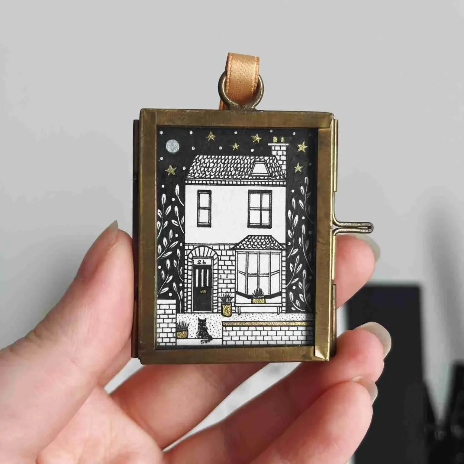 A small house illustration inside a metal frame with a leather strap, featuring a front door, windows, a bench, plants, and night sky with stars and a moon.