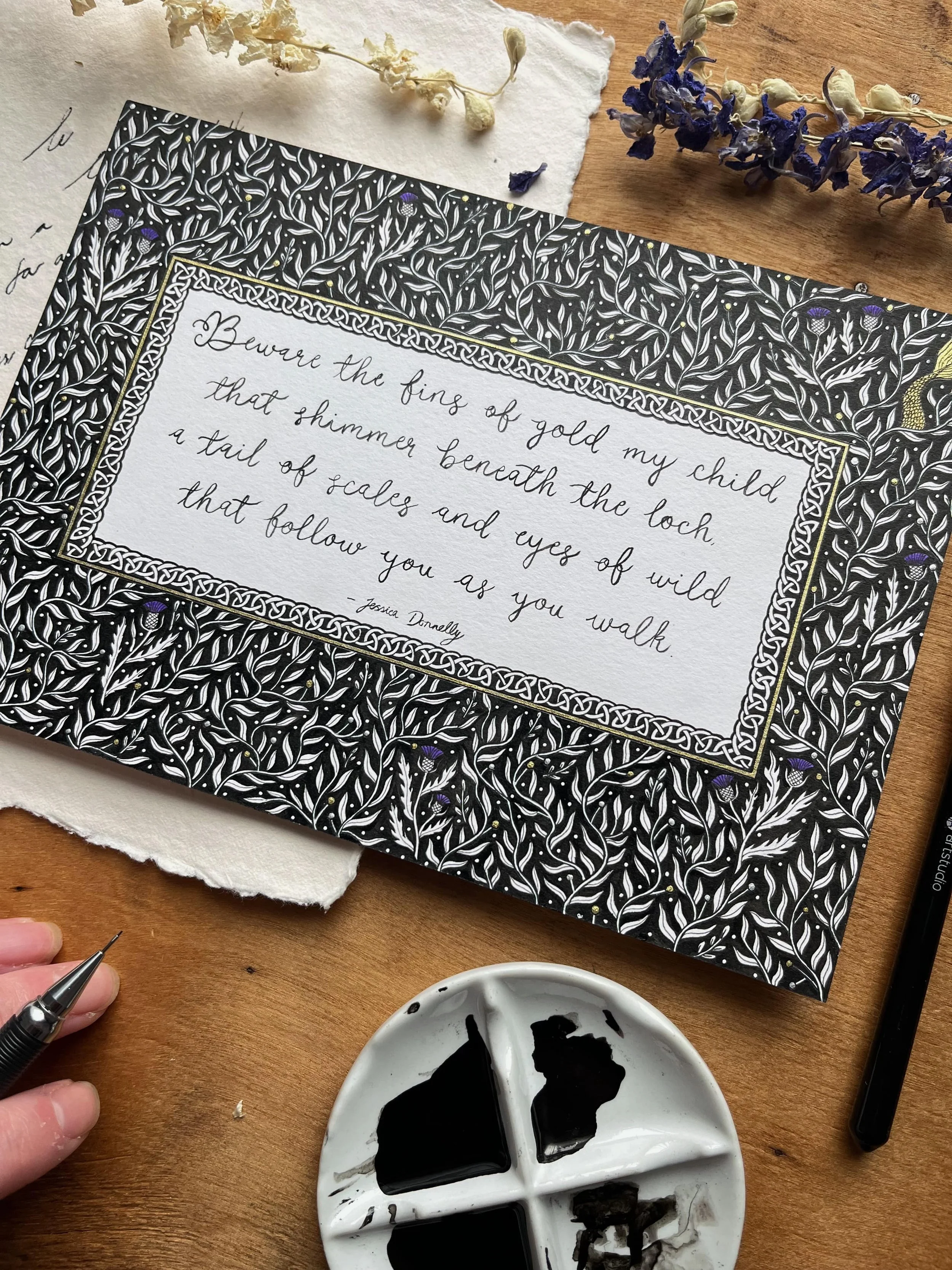 A decorative handwritten quote on a white card surrounded by a black and white floral patterned border: "Beware the sting of gold my child that shimmer beneath the loch a tail of scales and eyes of wild that follow you as you walk - Jessica Donnelly." The card is placed on a wooden surface with dried white and purple flowers, a black fine-liner pen, and a white palette with black ink splashes, and a person's hand holding a small tool.