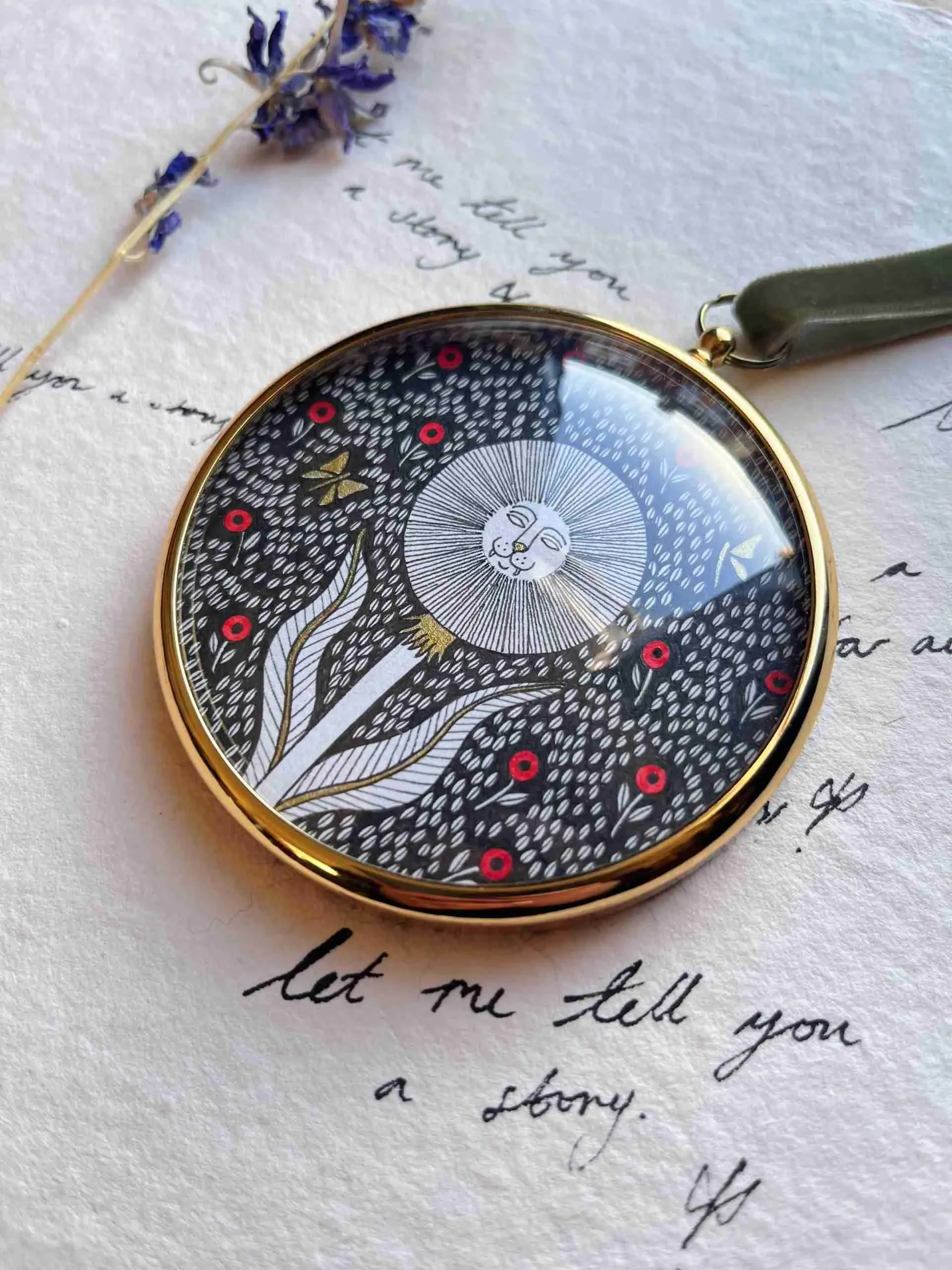 A round watch with a black and white lion illustration and red dots, placed on handwritten letter with a blue flower.