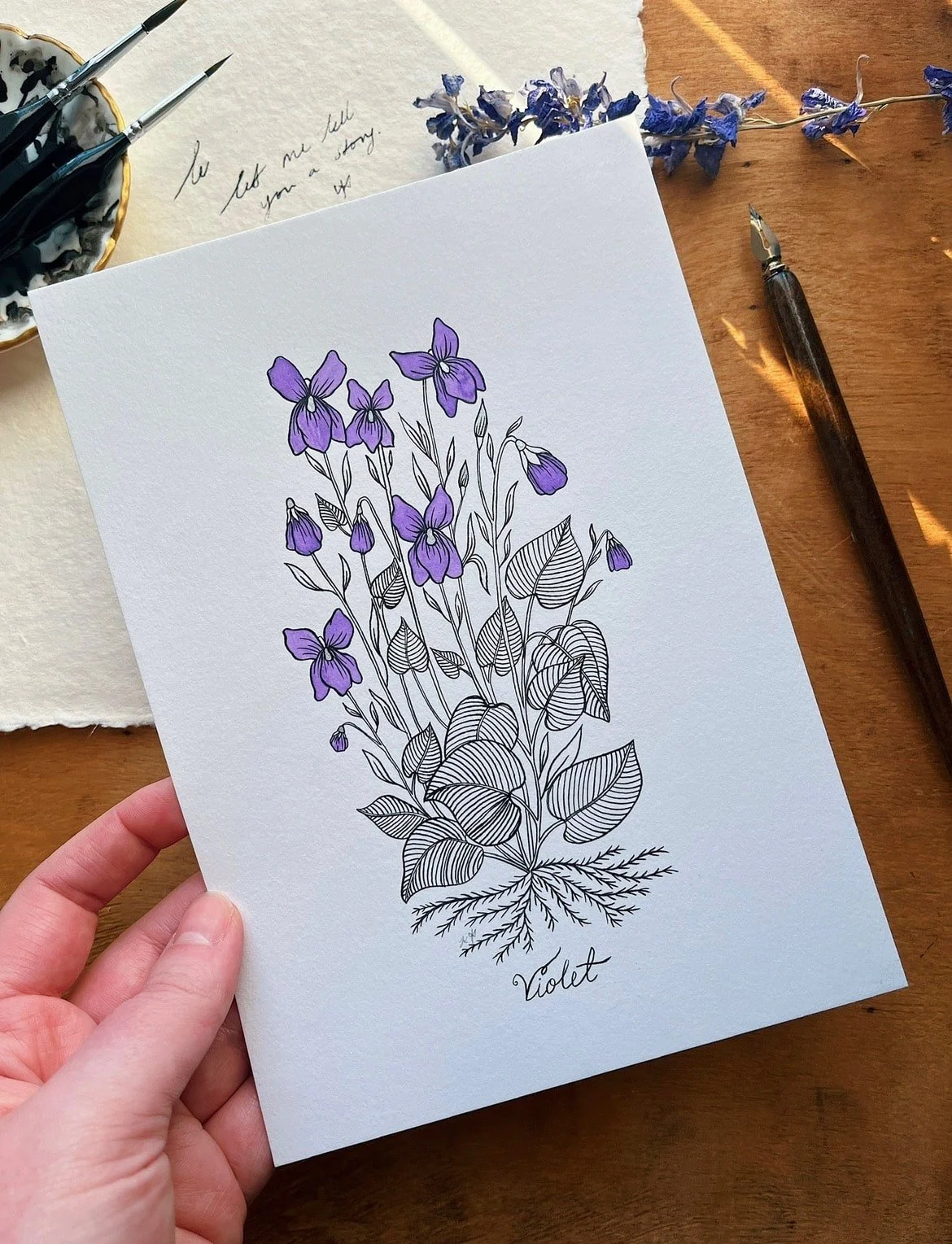 Hand holding a botanical illustration of violet flowers and leaves on white paper, labeled 'Violet', surrounded by art supplies and dried flowers on a wooden surface.