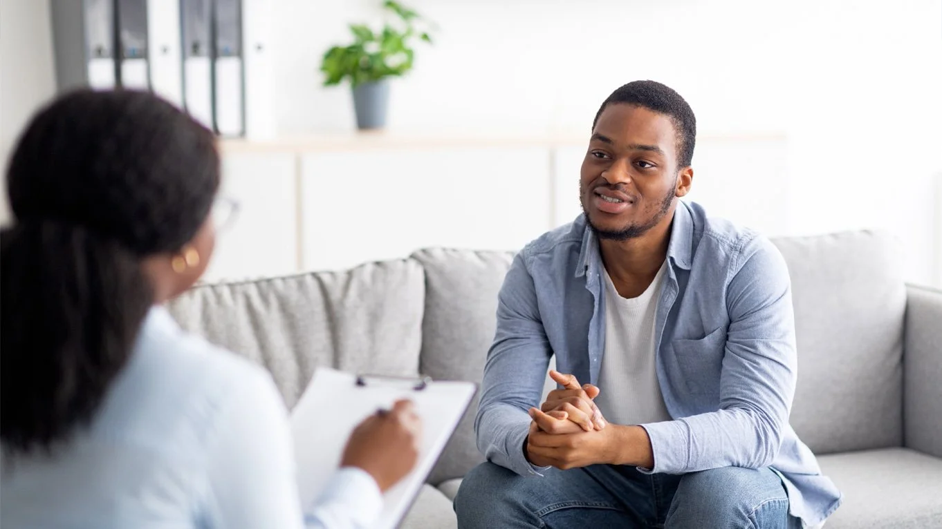 A man and woman sit across from each other. He sits on a couch and she takes notes