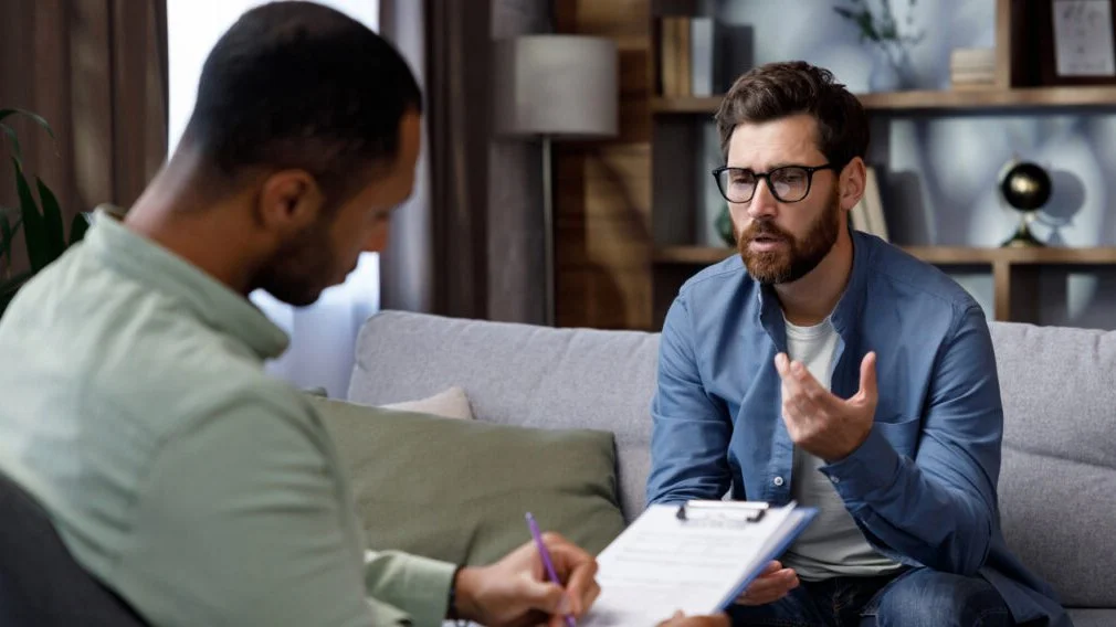 Two men sit across from each other and talk while one takes notes